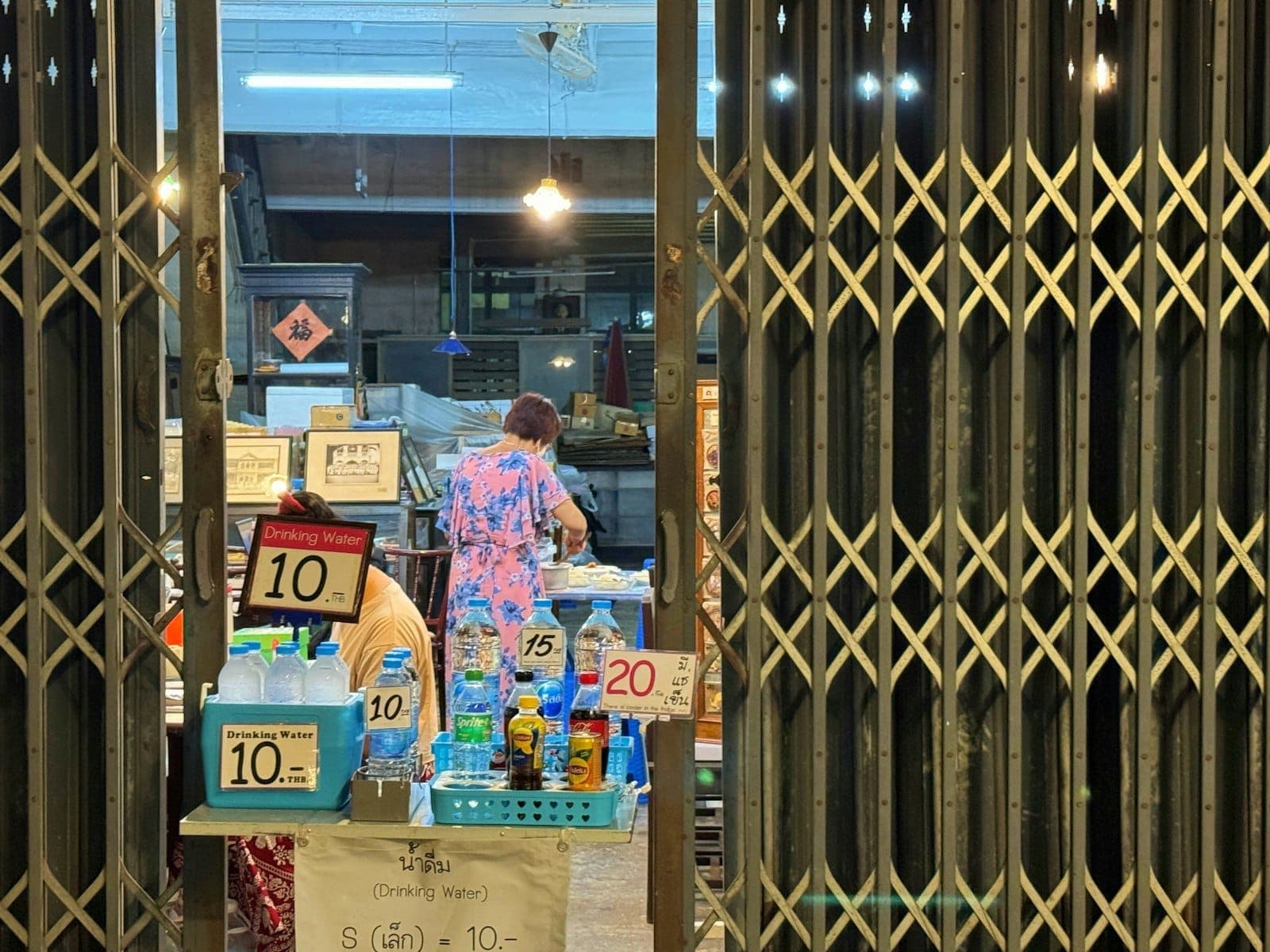 Panloob na tanawin ng isang maliit na tindahan ng pagkain sa Phuket na may mga inuming nakabalot sa bote na may presyong 10 hanggang 20 THB, isang babaeng naghahain ng pagkain, at isang lokal na kapaligiran.