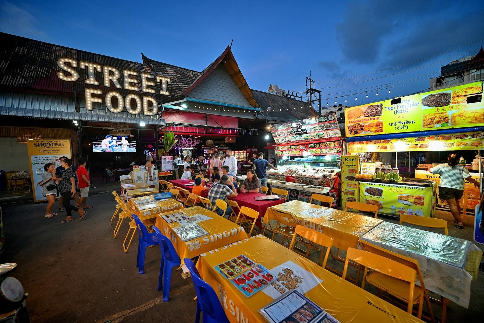 Outdoor na street food market sa Phuket sa takipsilim na may mga mesa, makulay na upuan, mga stall ng pagkain, at malaking tanda ng 'Street Food'.