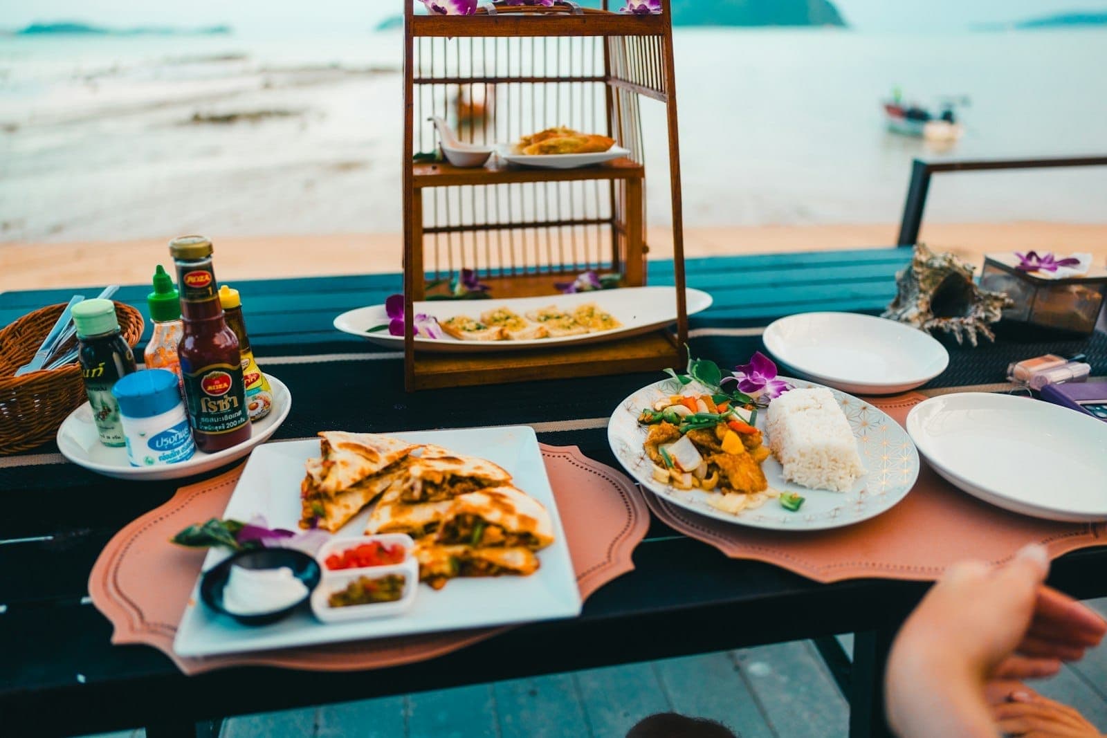 Isang mesa sa isang restaurant sa dalampasigan sa Phuket na may mga plato ng pagkaing Thai, mga sarsa, at mga inumin na may magandang tanawin ng karagatan.