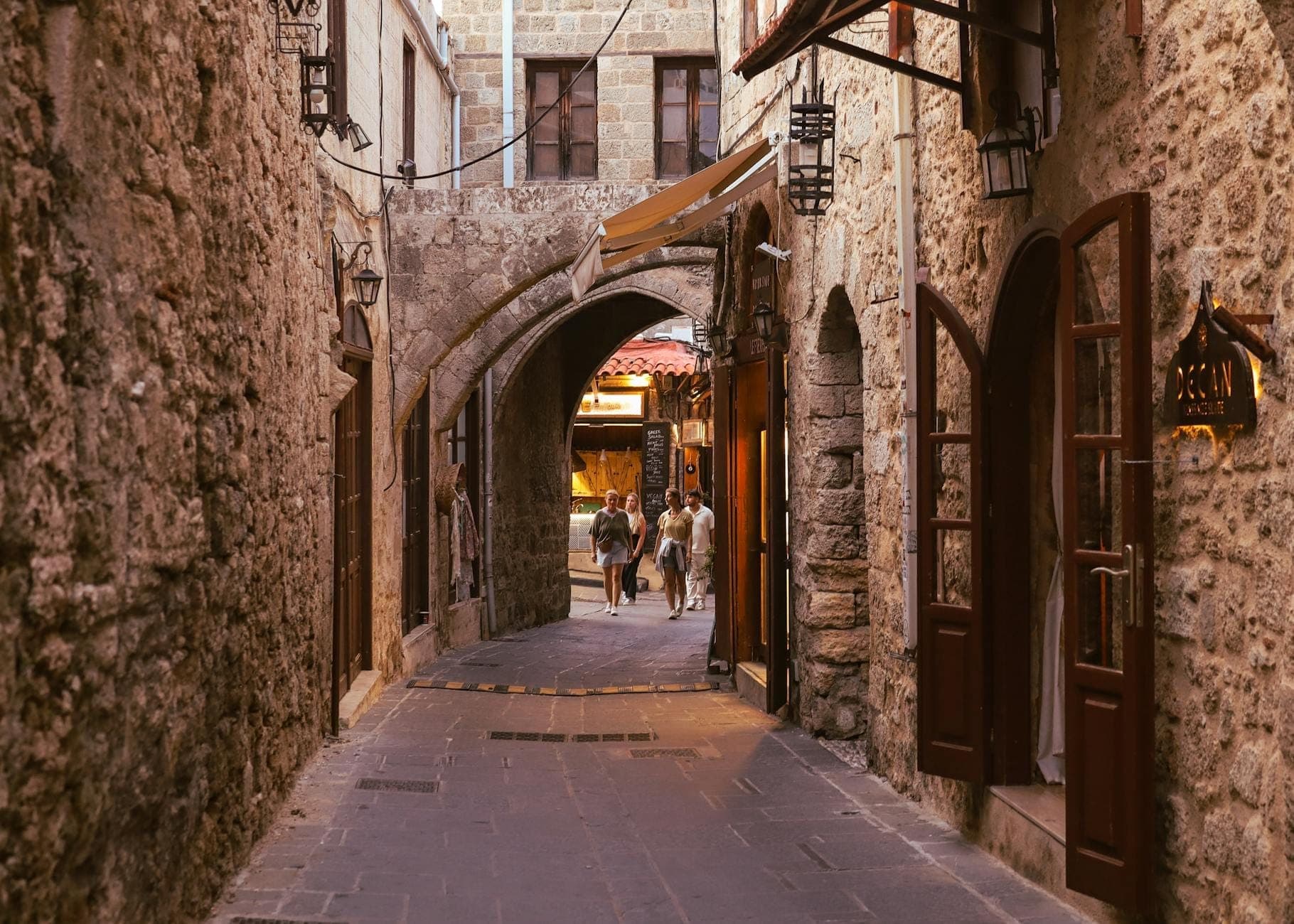 Charmante rue pavée dans la vieille ville de Rhodes avec des entrées de restaurants, une porte en arche et des personnes marchant en arrière-plan.