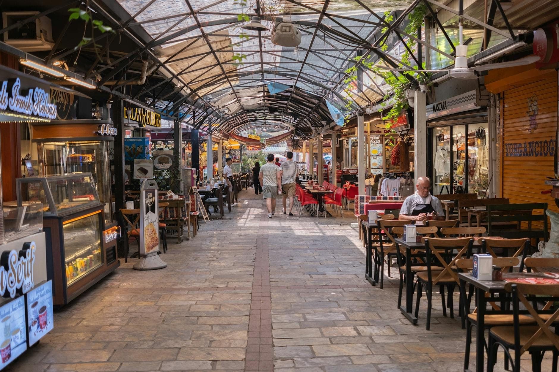Ruelle couverte bordée de cafés et boutiques décontractés, des personnes dînant à des tables simples, évoquant des restaurants locaux abordables.