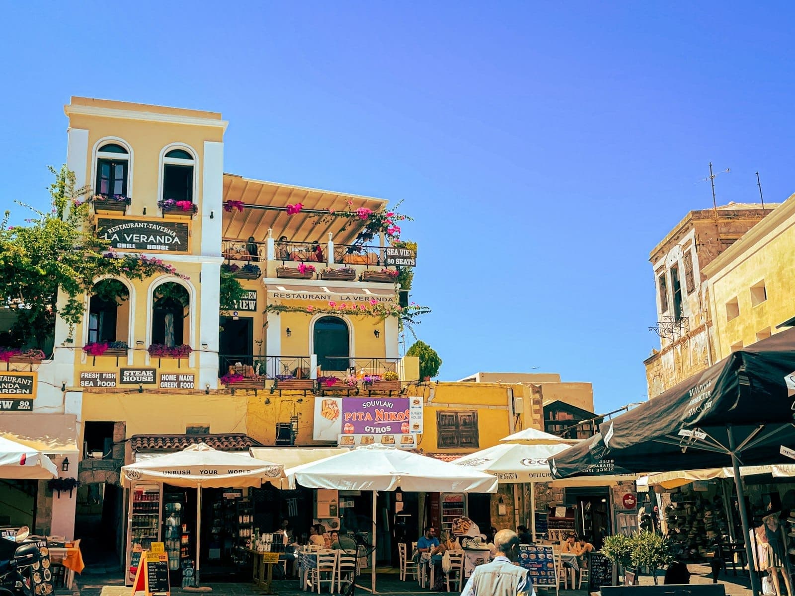 Restaurants et cafés en plein air avec des parasols colorés et des bâtiments méditerranéens historiques sur une place animée de la Vieille Ville de Rhodes sous un ciel bleu éclatant.