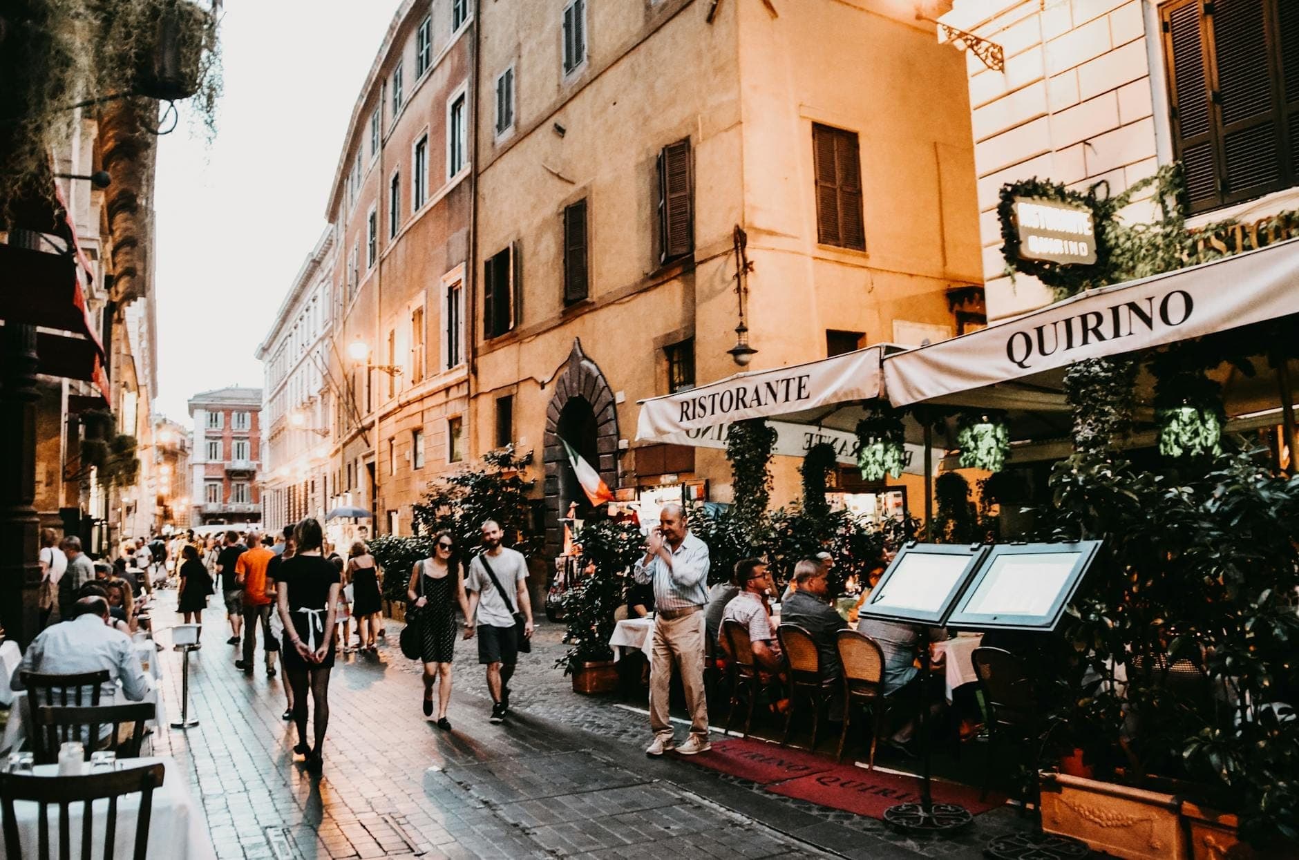 Belebte römische Straße mit Menschen, die im Freien in einem Ristorante speisen, Fußgängern und historischen Gebäuden im Hintergrund in der Abenddämmerung.