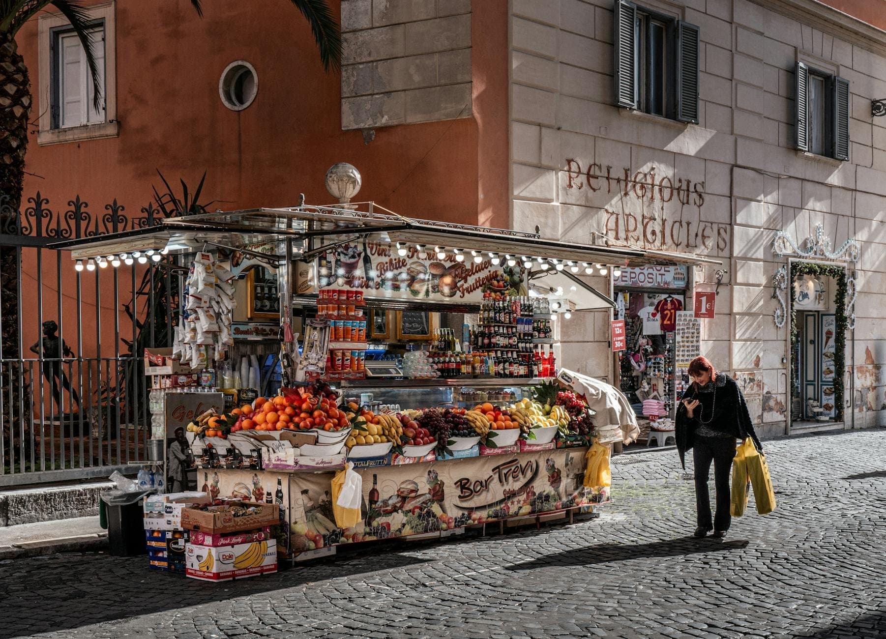 Ein Straßenimbiss- und Marktstand in Rom, der Obst, Snacks und Getränke verkauft, vor einem klassischen römischen Gebäude mit vorbeigehenden Menschen.