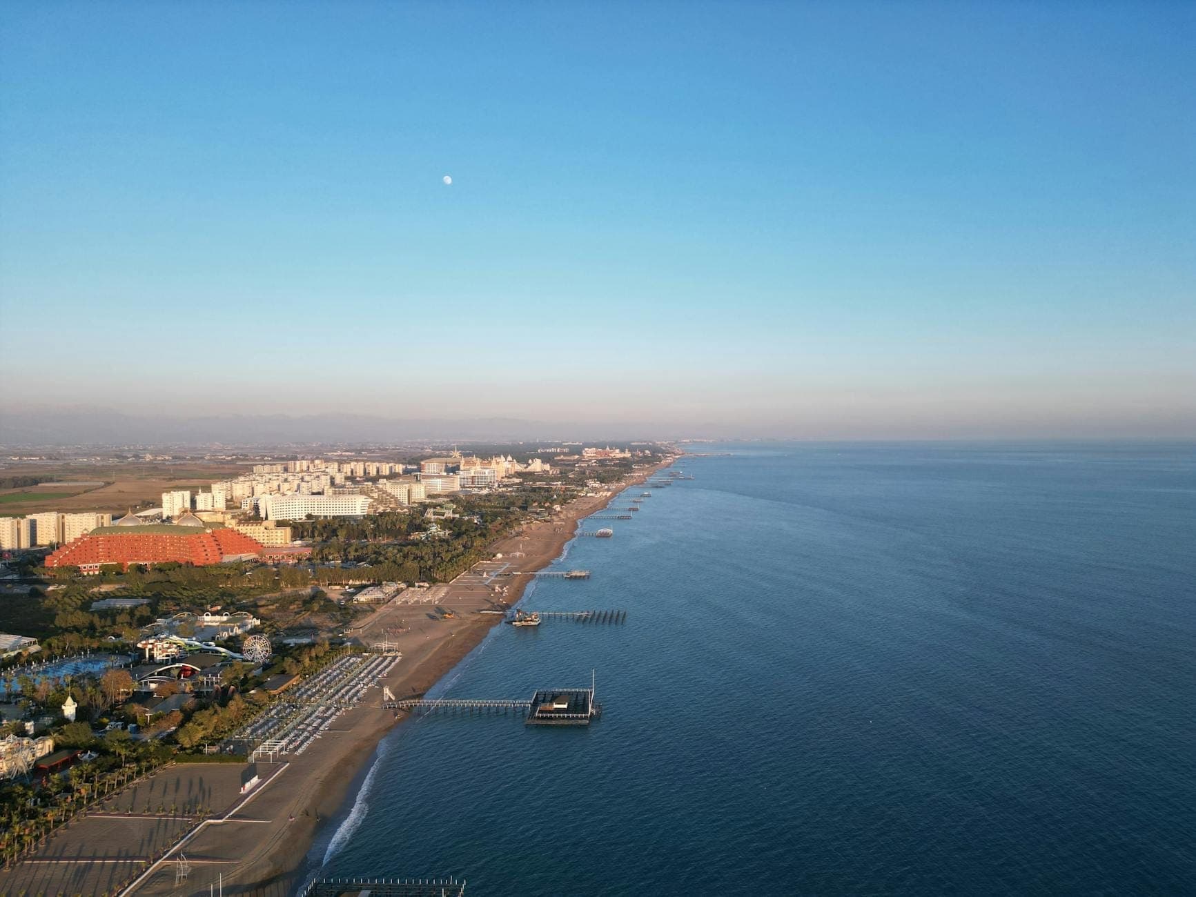 Vue aérienne de Lara Beach avec un long littoral de sable, des eaux calmes et claires, et des rangées de grands hôtels de villégiature le long de la côte.