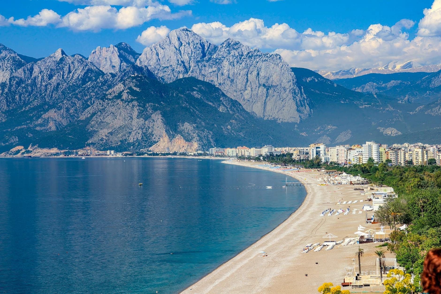 Vue panoramique de la plage de Konyaaltı avec le paysage urbain, le rivage de galets, l'eau bleue cristalline et les impressionnantes montagnes Taurus en arrière-plan.