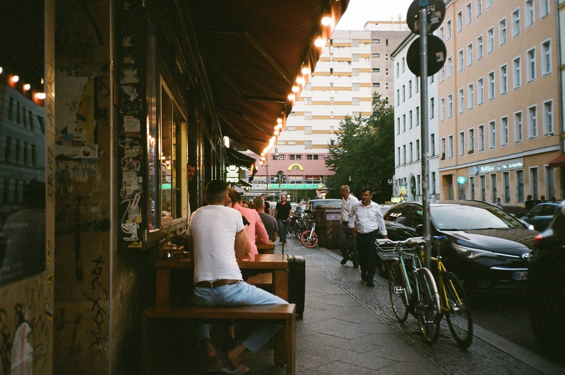 Cena movimentada na calçada com pessoas sentadas em mesas ao ar livre, bicicletas, prédios de apartamentos e vida urbana, típica dos bairros modernos de Berlim.
