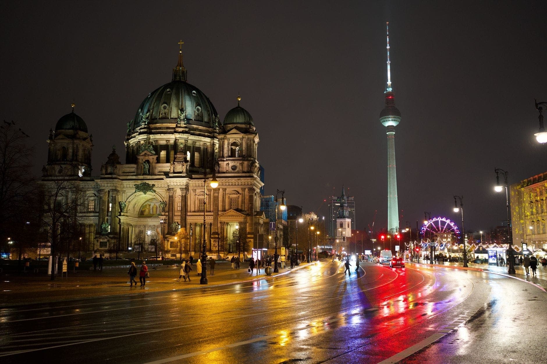 Vista noturna da Catedral de Berlim e da torre de televisão Fernsehturm, com luzes refletidas nas ruas molhadas e pessoas caminhando por perto.