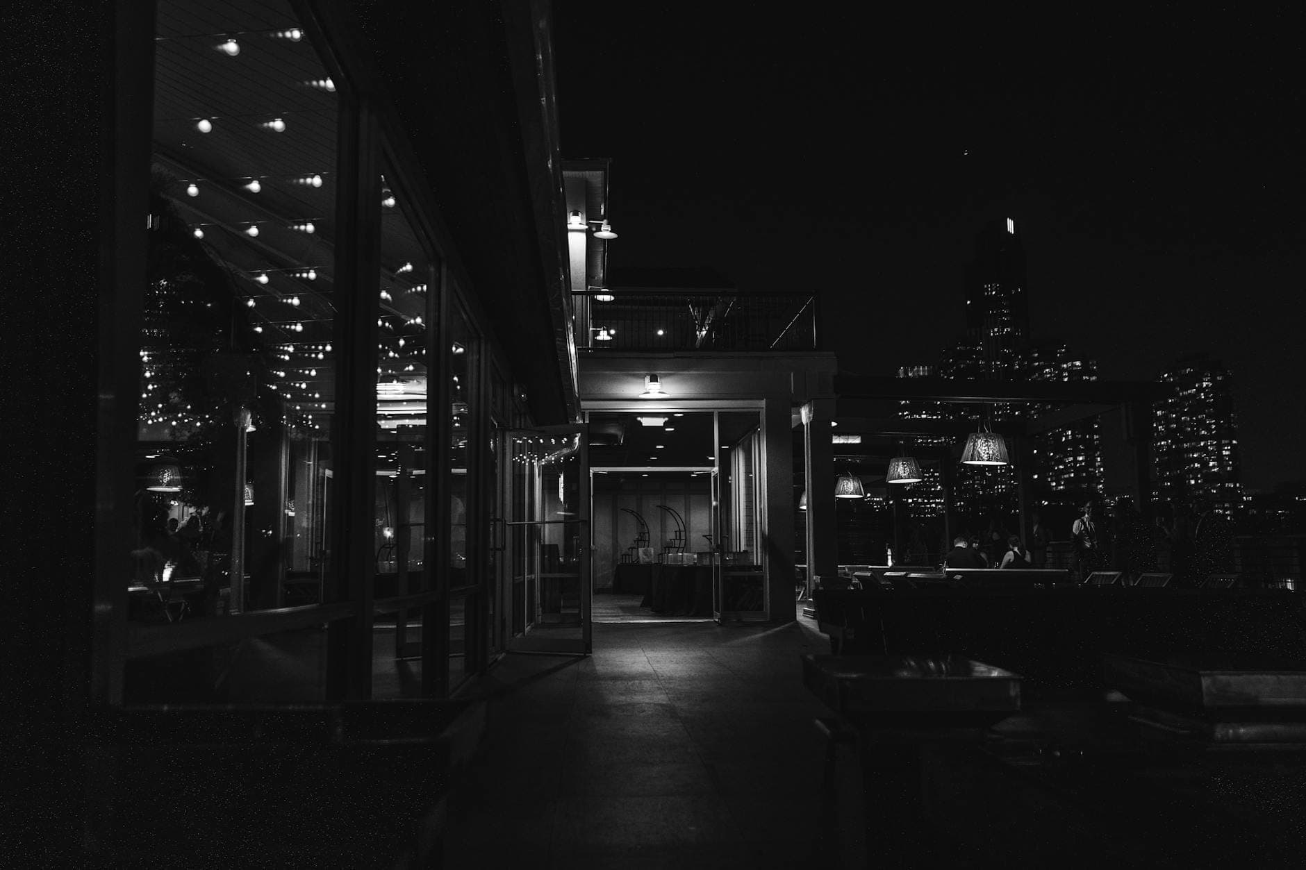 Black and white photo of a rooftop bar or restaurant at night with city lights in the background.