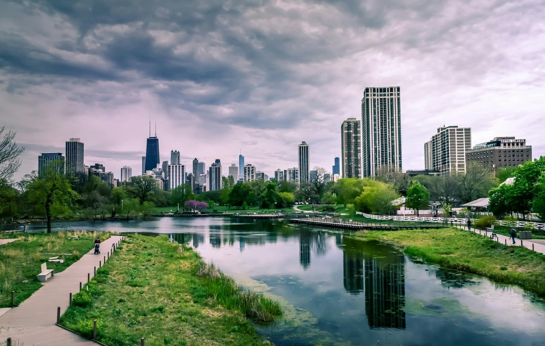 Chicago skyline behind Lincoln Park’s pond and walking paths under a dramatic cloudy sky, showing green landscape and cityscape together.