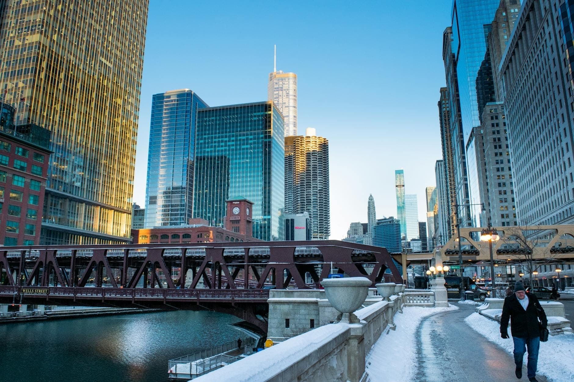 Winter scene in downtown Chicago with snow on sidewalks, Chicago River, bridges, and modern highrise buildings under clear blue sky.