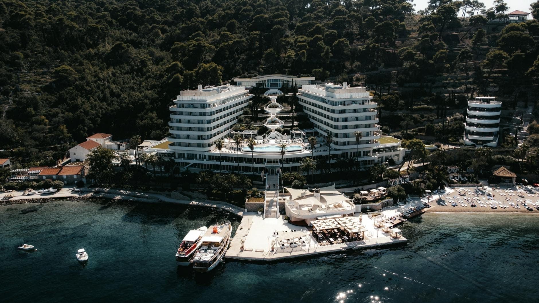 Pemandangan udara hotel resor tepi pantai modern dengan kolam renang dan akses langsung ke laut, dikelilingi pepohonan hijau yang rimbun di pesisir Dubrovnik.