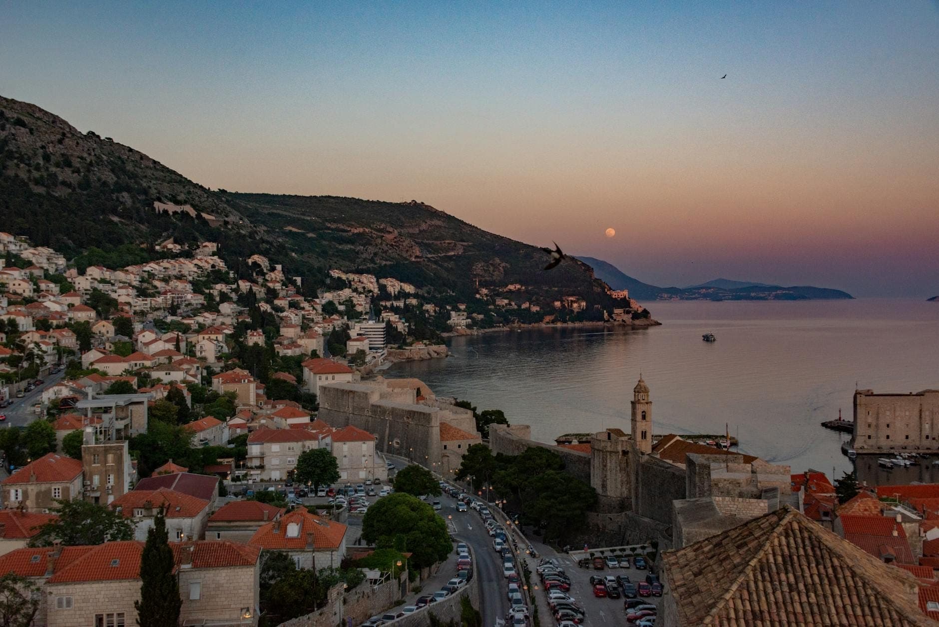 Pemandangan dari ketinggian atas Dubrovnik saat senja menampilkan jalan-jalan kota, tembok Kota Tua, rumah-rumah di lereng bukit, dan garis pantai Adriatik.
