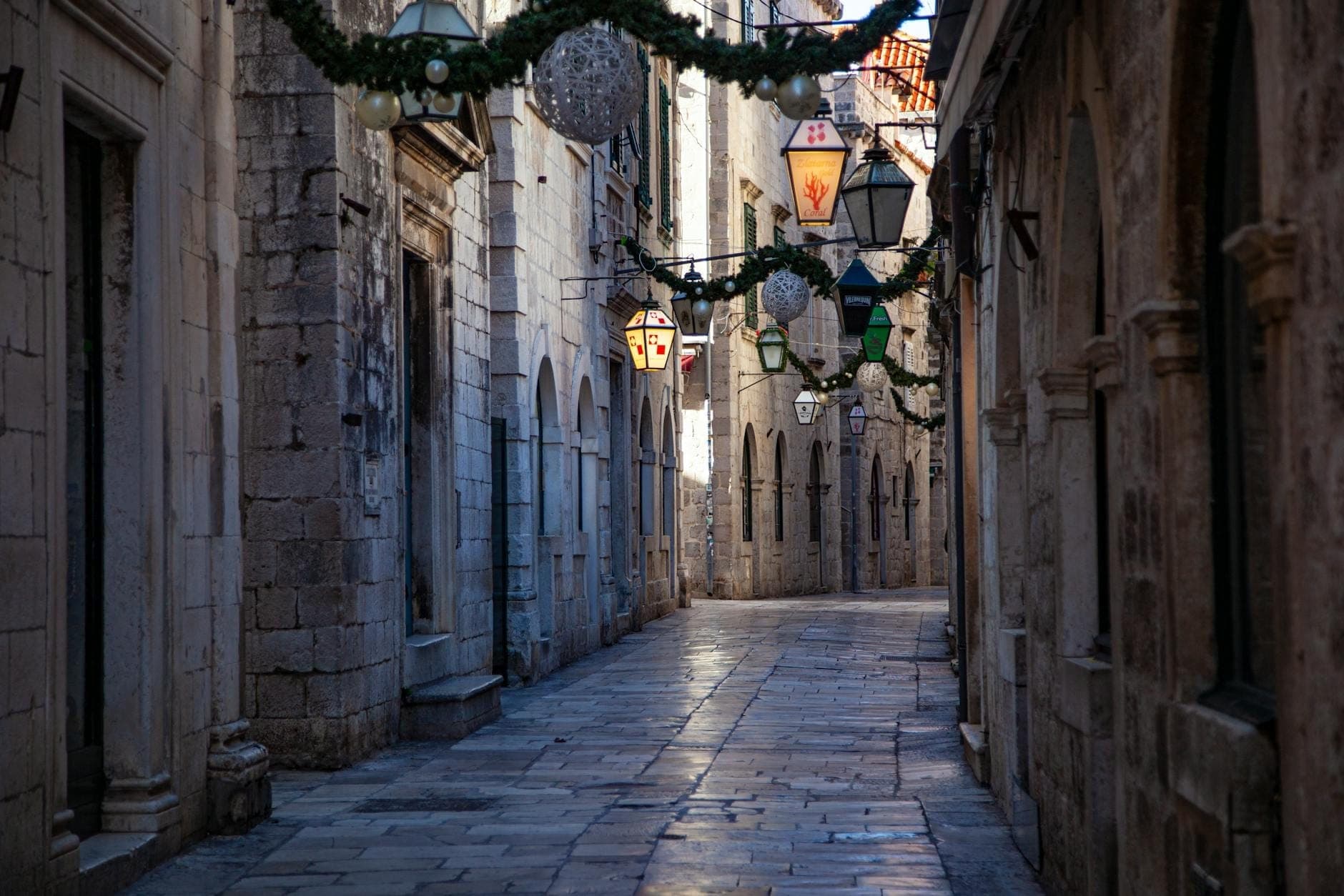 Jalan batu sempit dan kosong di Kota Tua Dubrovnik dengan lentera dan ornamen yang digantung di atas. Bangunan bersejarah dari batu kapur berjajar di kedua sisi.