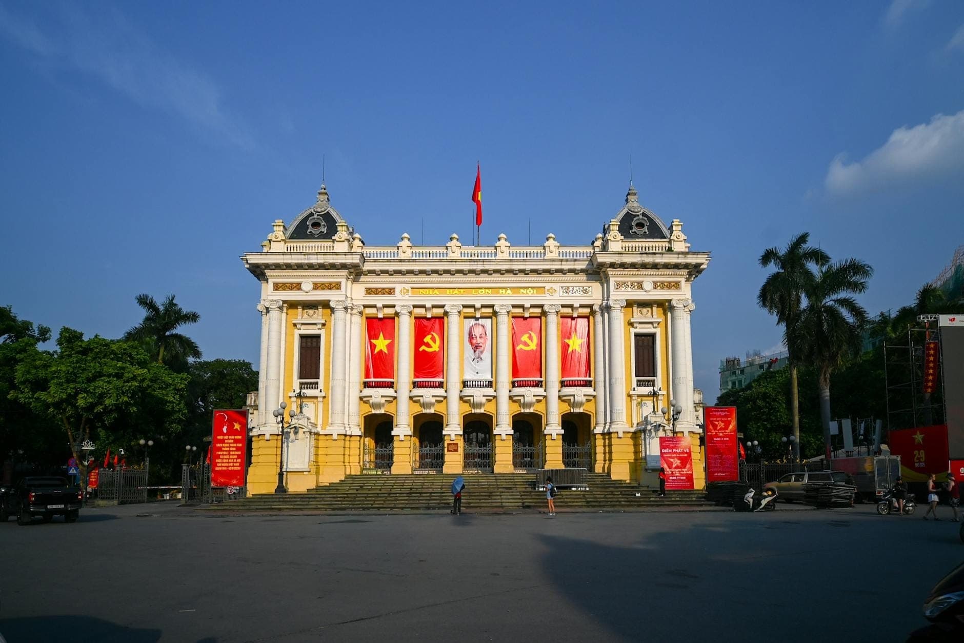Vista frontal del Hanoi Opera House con arquitectura colonial francesa, grandes columnas y banderas vietnamitas en una amplia plaza iluminada por el sol.
