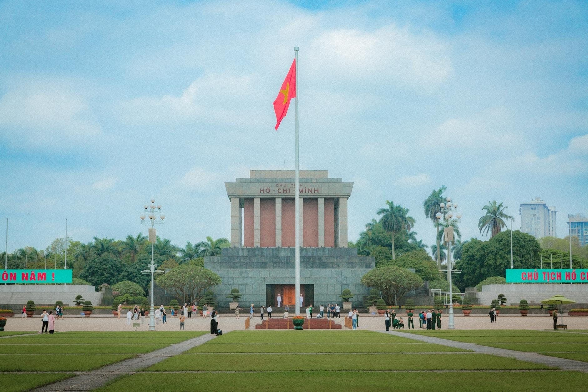 Vista diurna amplia del Mausoleo de Ho Chi Minh en la Plaza Ba Dinh, Hanói, con una bandera vietnamita, céspedes verdes y visitantes caminando cerca.