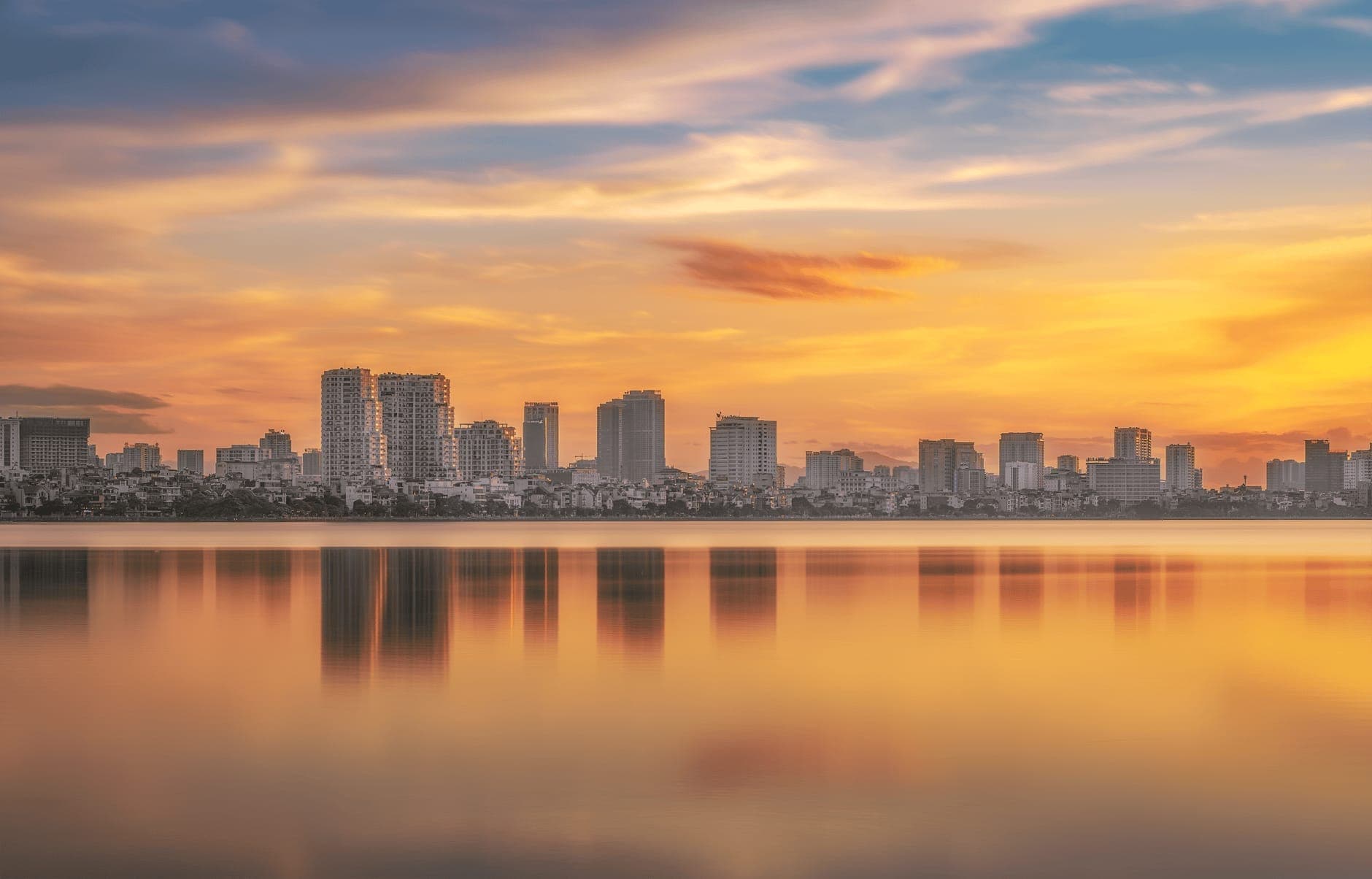 Vista panorámica amplia del Lago del Oeste en Hanói al atardecer, con edificios modernos y un cielo colorido reflejado en el agua tranquila.