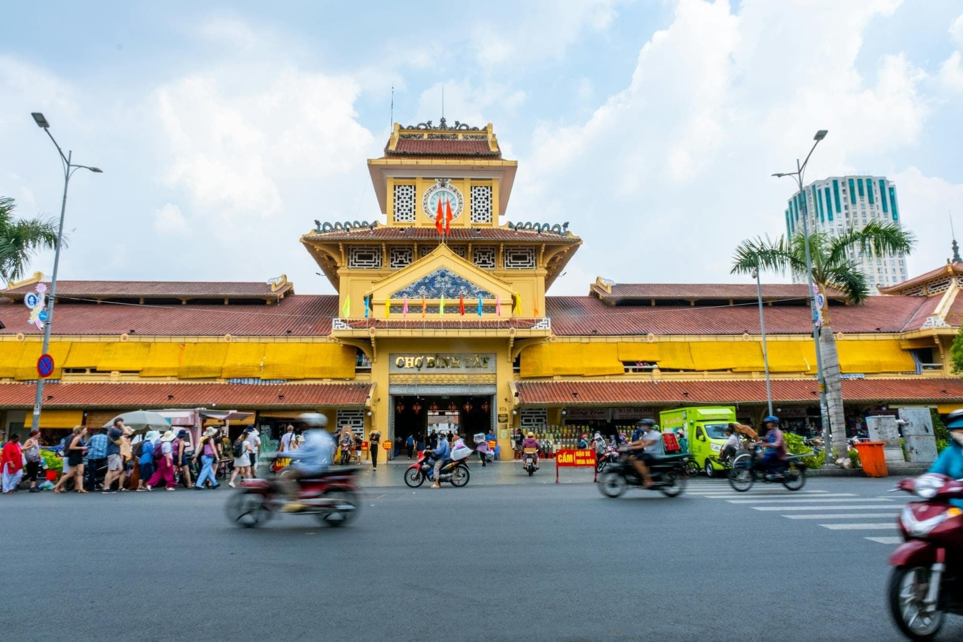 Malawak na tingin sa malaking dilaw na gusali ng palengke ng Chợ Bình Tây, maraming tao at motorsiklo sa labas, sa Chợ Lớn, Lungsod ng Ho Chi Minh.