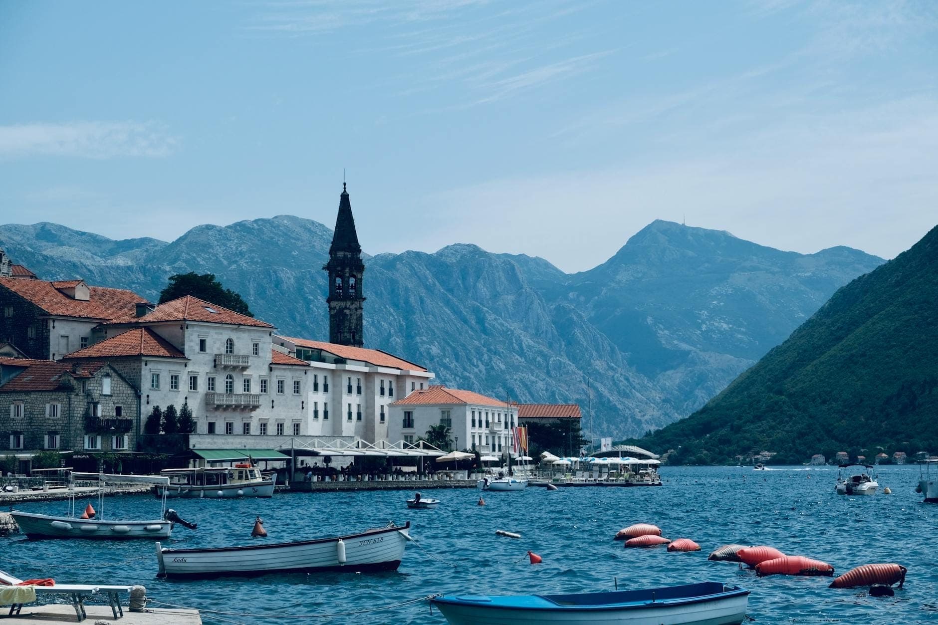 Des bâtiments médiévaux en pierre aux toits rouges longent le front de mer de Kotor, au Monténégro, avec des bateaux flottant sur la baie et des montagnes spectaculaires en arrière-plan.