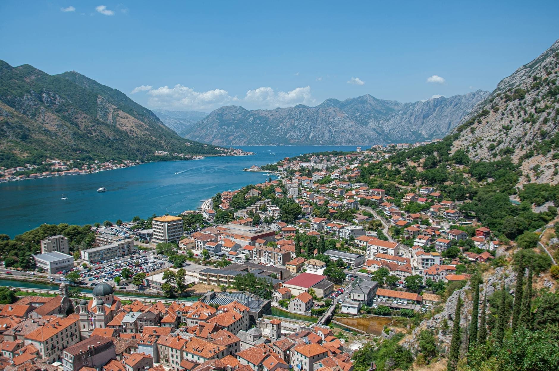 Vue panoramique de la baie et de la ville de Kotor, entourée de montagnes avec des toits de tuiles rouges et une eau bleue visible sous un ciel dégagé.