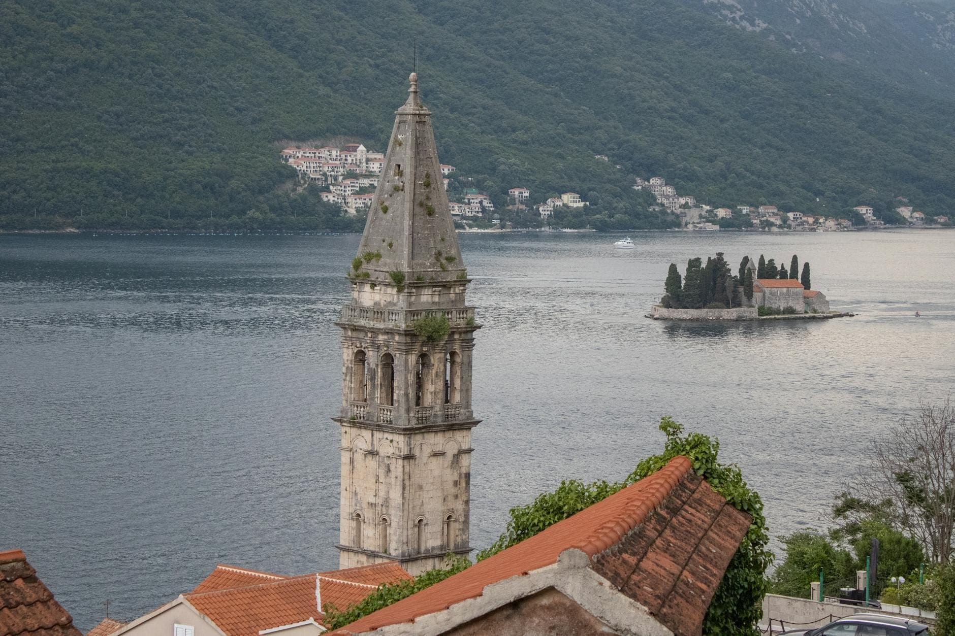 Vue du clocher et des toits de Perast au premier plan avec l'île de Saint-Georges et les montagnes de l'autre côté de la Baie de Kotor en arrière-plan.
