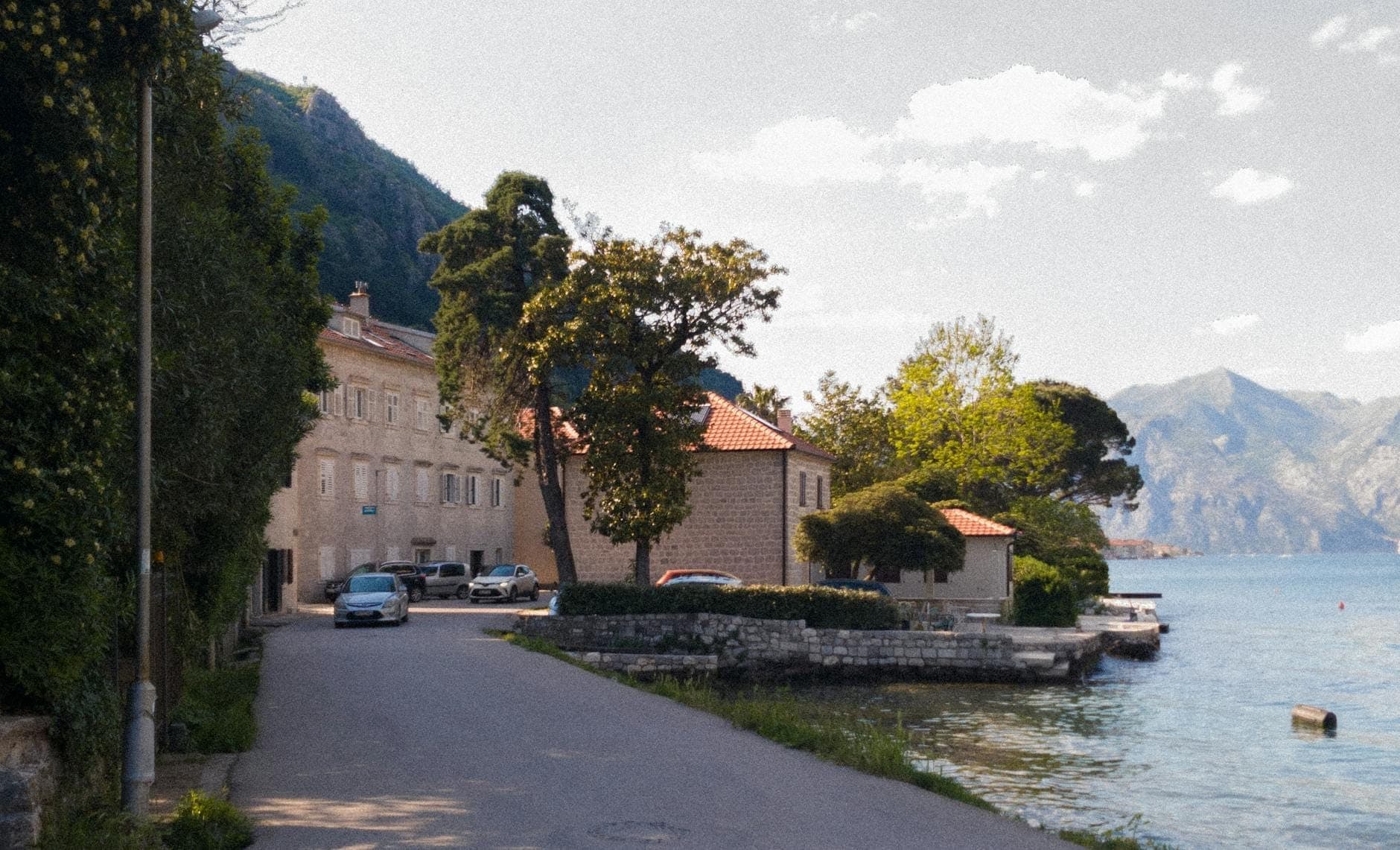 Route pittoresque en bord de baie dans un village tranquille près de Kotor, avec des maisons en pierre, des montagnes et une eau calme.