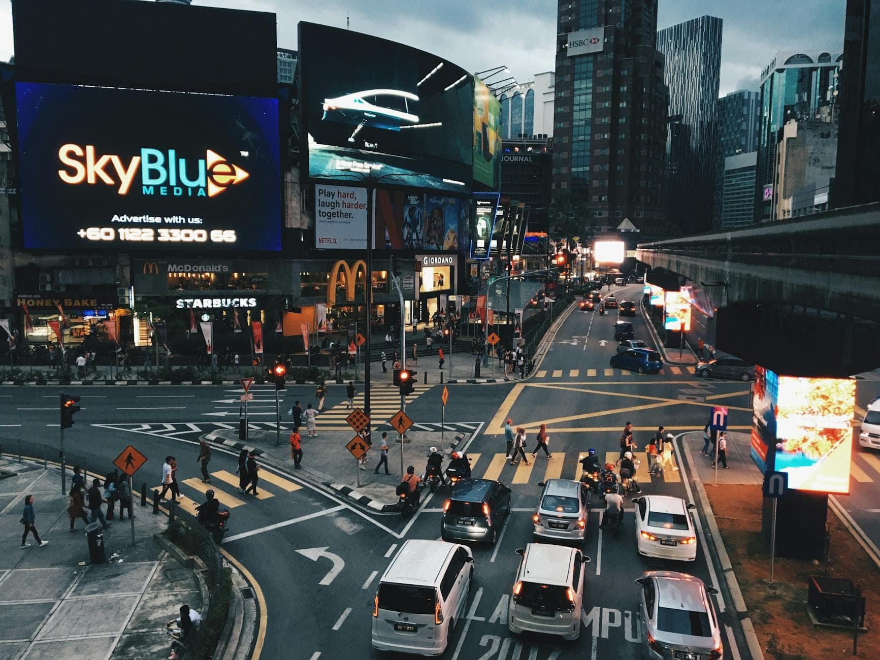 Busy intersection in Bukit Bintang, Kuala Lumpur, with cars, pedestrians, and brightly lit billboards above McDonald's and Starbucks in the evening.