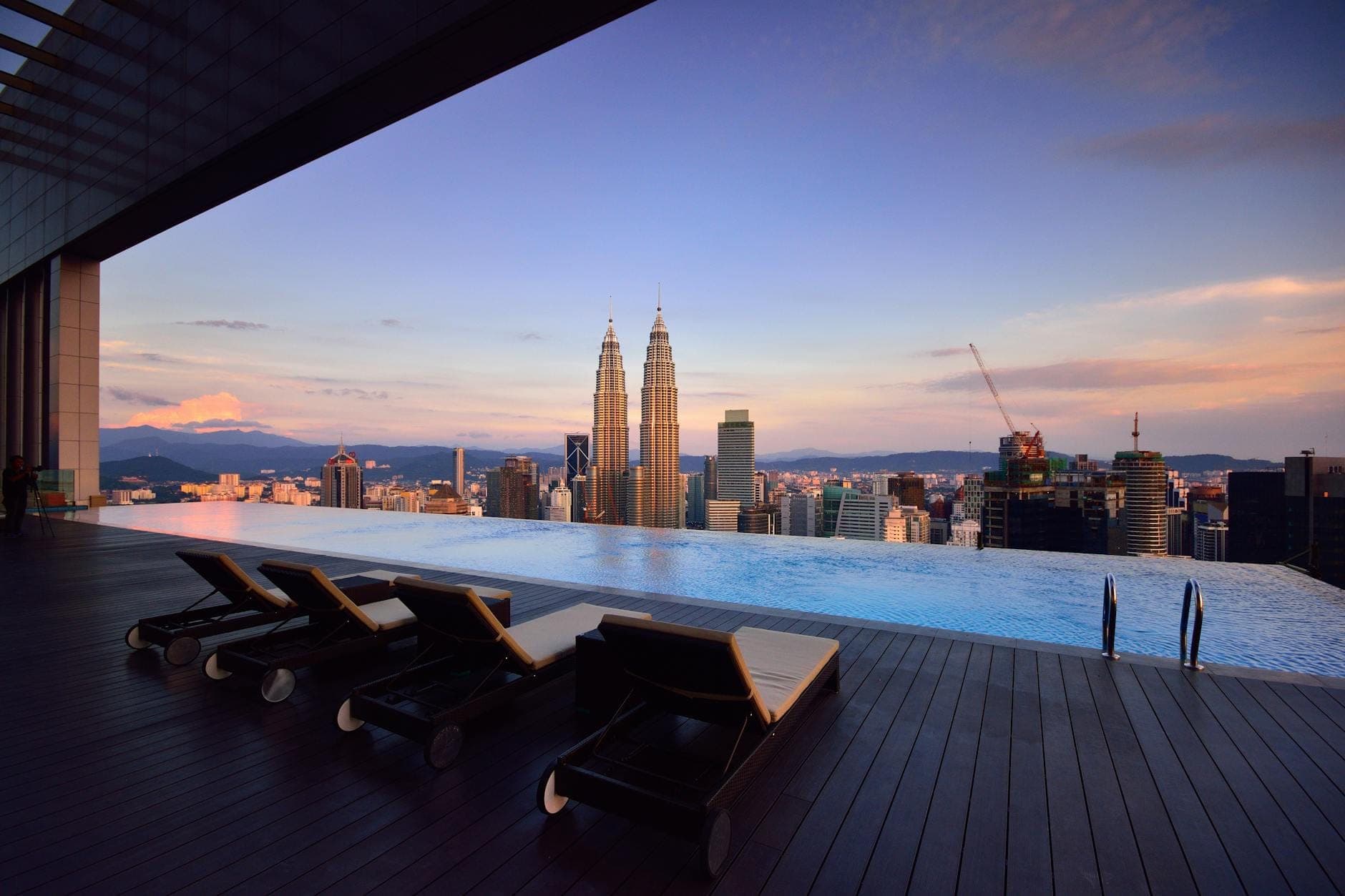 Infinity pool with lounge chairs overlooking Kuala Lumpur skyline and Petronas Twin Towers at sunset, featuring warm city lights and modern buildings.