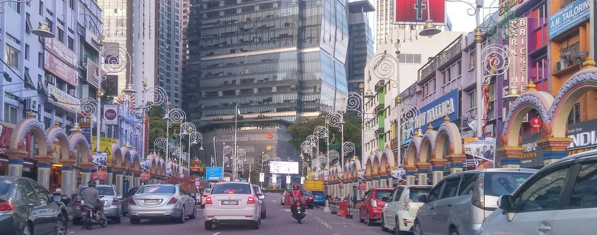 Street view of Kuala Lumpur’s vibrant Brickfields district, featuring colorful archways, busy traffic, and modern skyscrapers rising in the background.