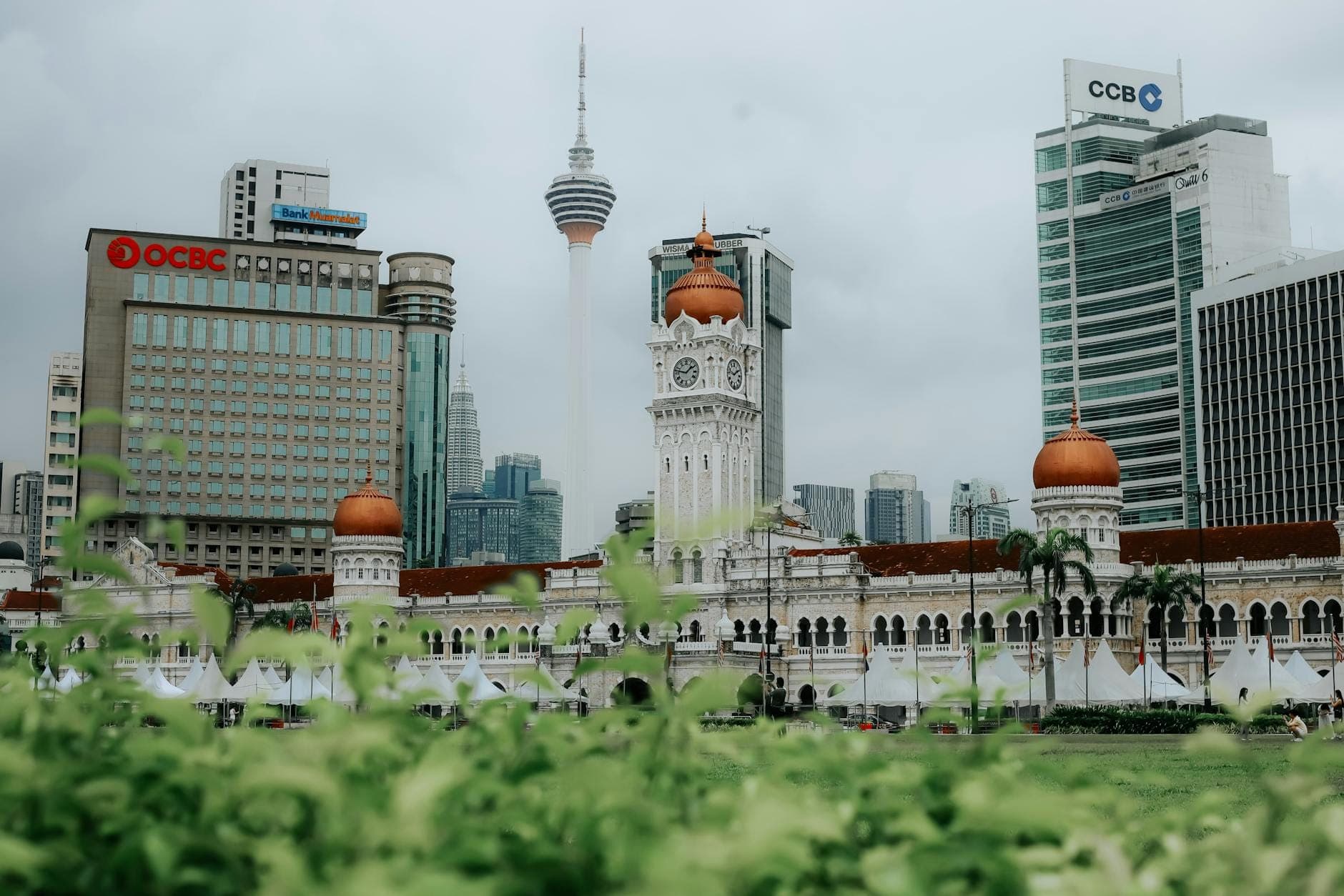 View of Sultan Abdul Samad Building with its iconic clock tower and copper domes, surrounded by Kuala Lumpur skyscrapers on a cloudy day.