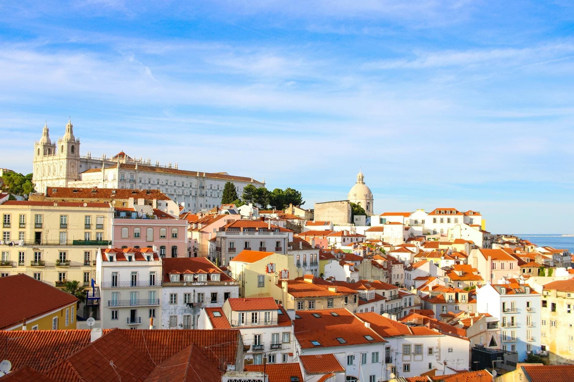 Vista panorâmica e ensolarada do icônico bairro da Alfama, em Lisboa, com telhados de cerâmica, casarões históricos brancos, céu azul e pontos turísticos ao fundo.