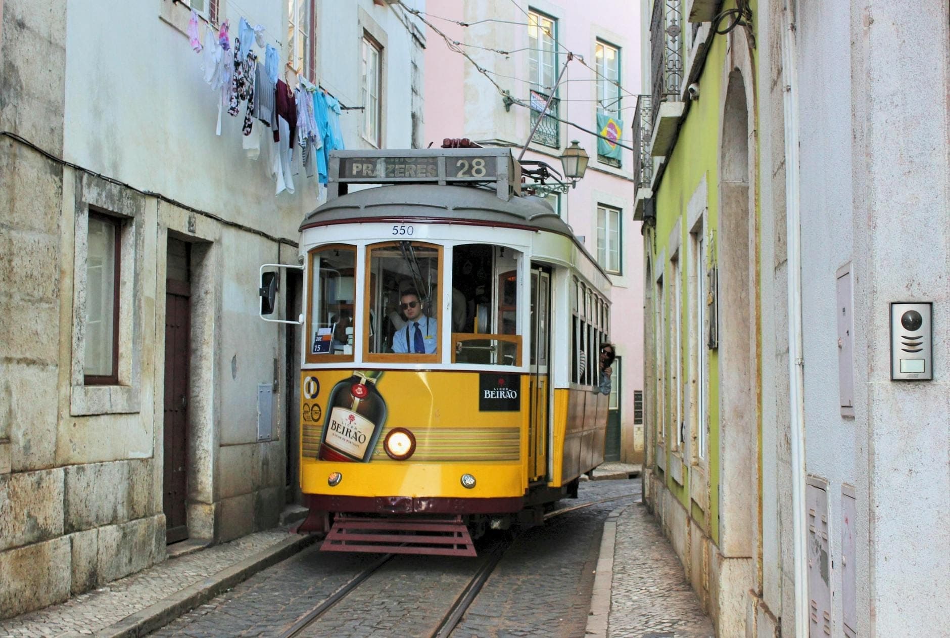 Um clássico elétrico amarelo passa por uma rua estreita e de paralelepípedos ladeada por velhos edifícios caiados e roupa estendida em Alfama, Lisboa.