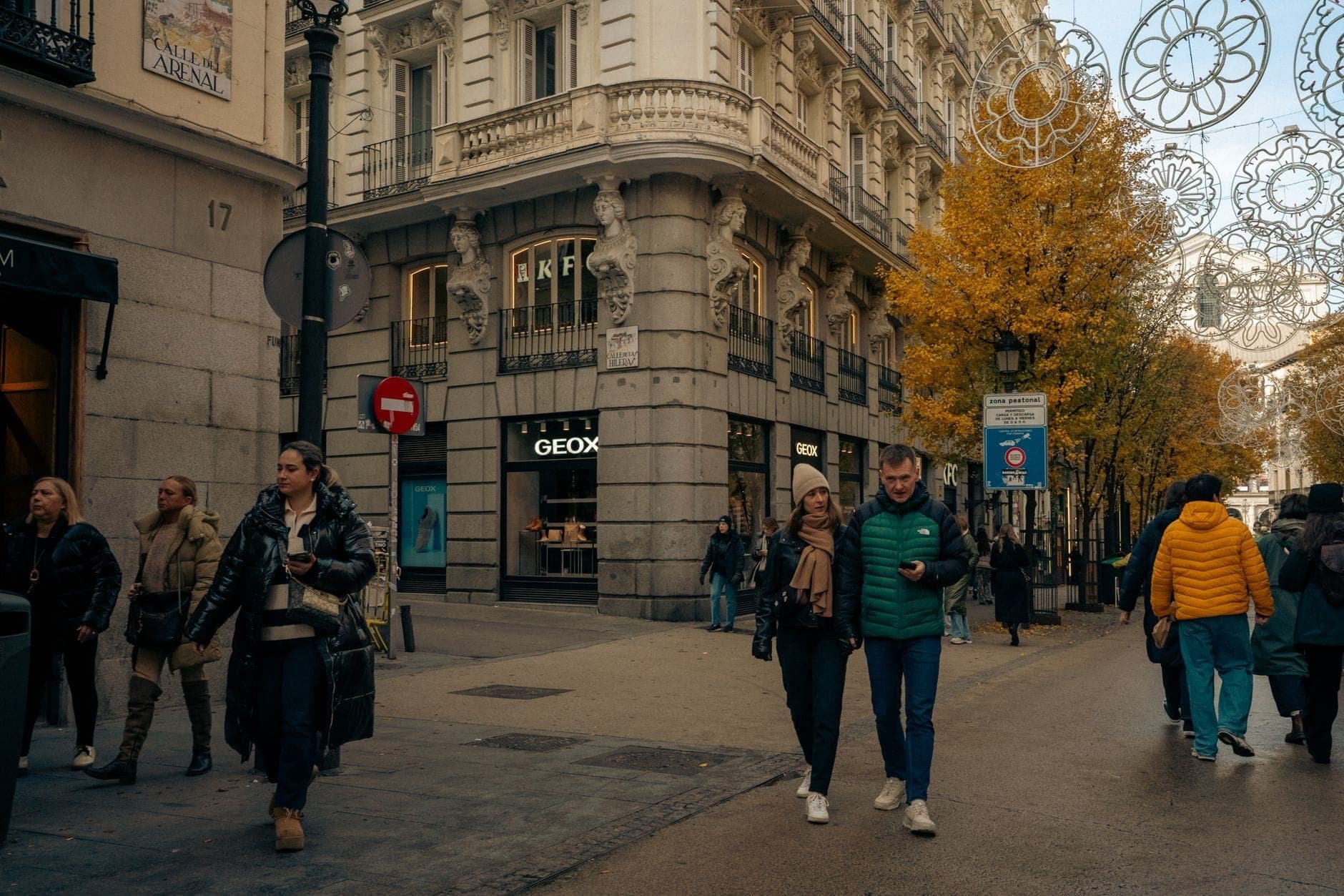 Elegante scena di strada a Madrid con ornati edifici di fine Ottocento, negozi di lusso e persone che camminano su un ampio viale fiancheggiato da alberi autunnali.
