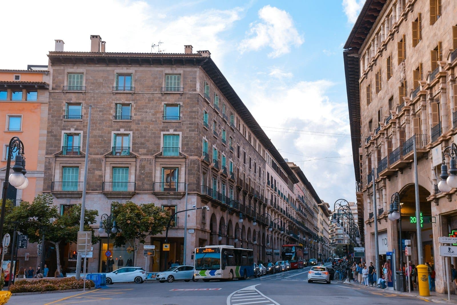 Breite Straßenszene im Zentrum von Palma de Mallorca mit historischen Steingebäuden, Bogenbalkonen, Stadtbussen, geparkten Autos und Fußgängern unter einem hellen Himmel.