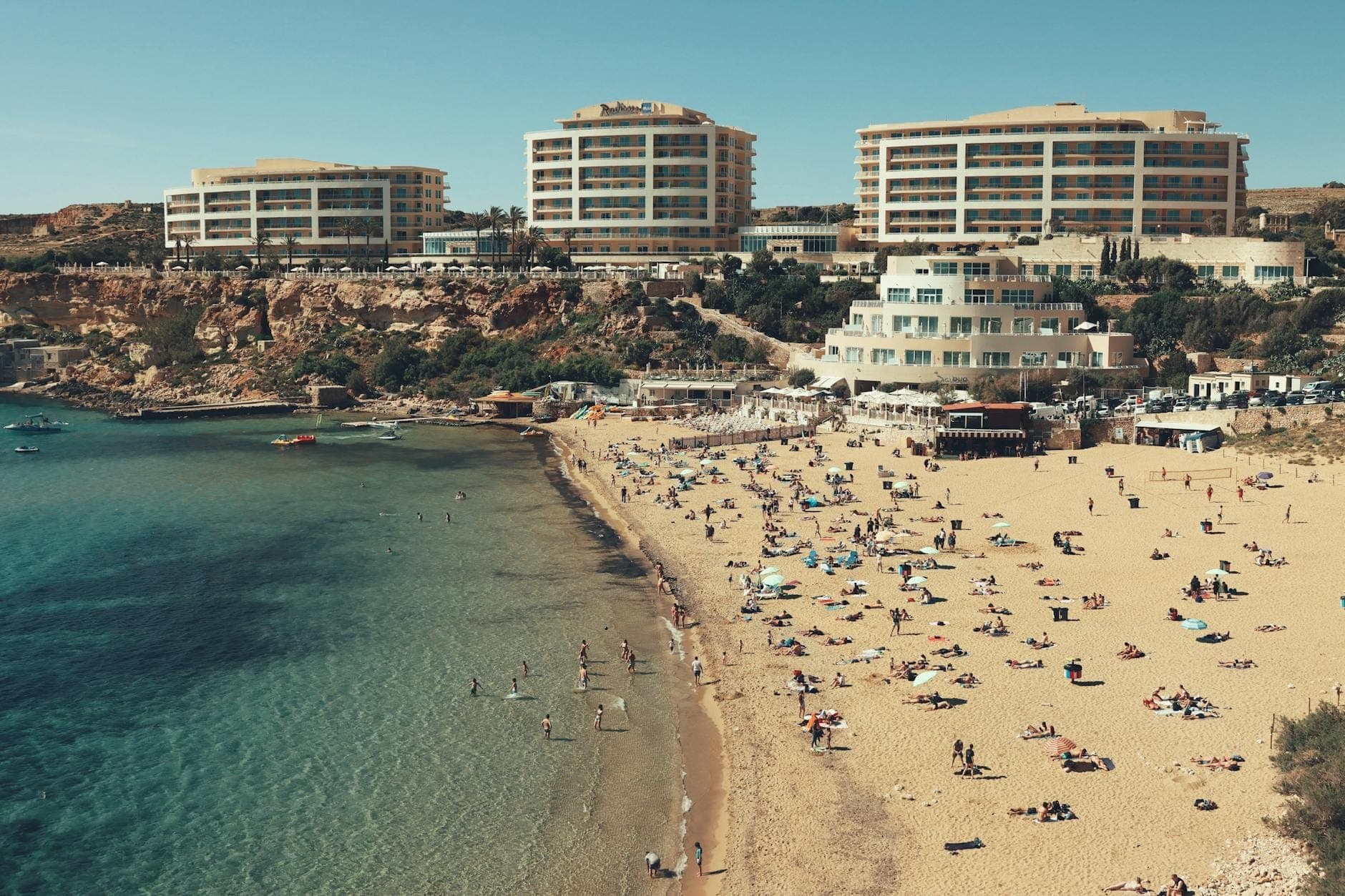 Großer Sandstrand mit Sonnenbadenden und Schwimmern, im Hintergrund moderne Hotels auf einem Höhenrücken, klares blaues Meer im Vordergrund.