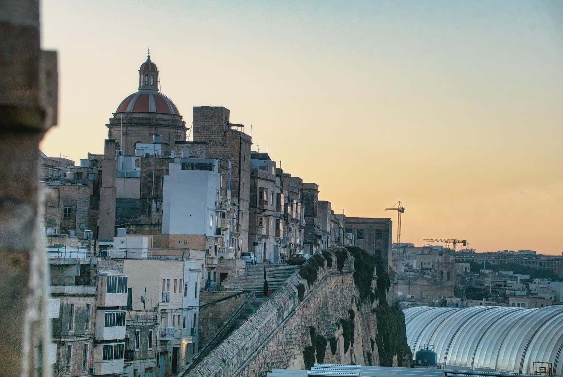 Blick auf die Stadtlandschaft von Valletta bei Sonnenuntergang mit Steingebäuden, Kuppelkirche und historischer Festungsmauer.