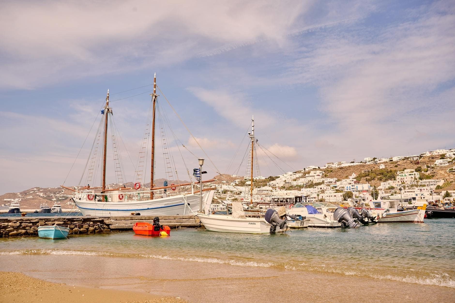 Boote, die in einer ruhigen, seichten Bucht nahe einem Sandstrand ankern, mit weiß getünchten Gebäuden auf einem Hügel im Hintergrund unter einem sonnigen Himmel.