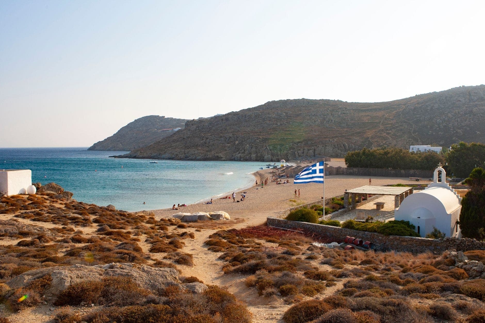 Eine ruhige Sandbucht mit wenigen Menschen, einer griechischen Flagge, einer kleinen weißen Kapelle und Hügeln im Hintergrund in Mykonos.