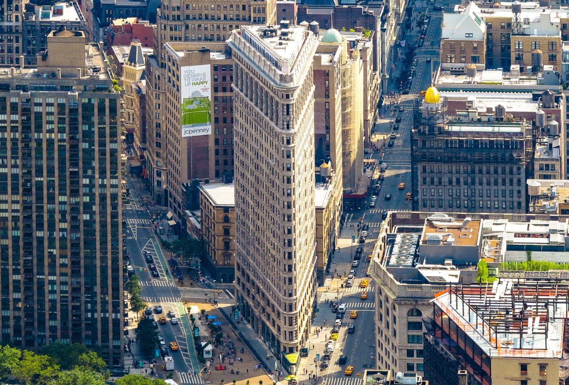 มุมมองทางอากาศของ Flatiron Building และถนนโดยรอบในแมนฮัตตันท่ามกลางแสงแดดจ้า แสดงให้เห็นสถาปัตยกรรมสามเหลี่ยมอันเป็นเอกลักษณ์และความคึกคักของเมือง