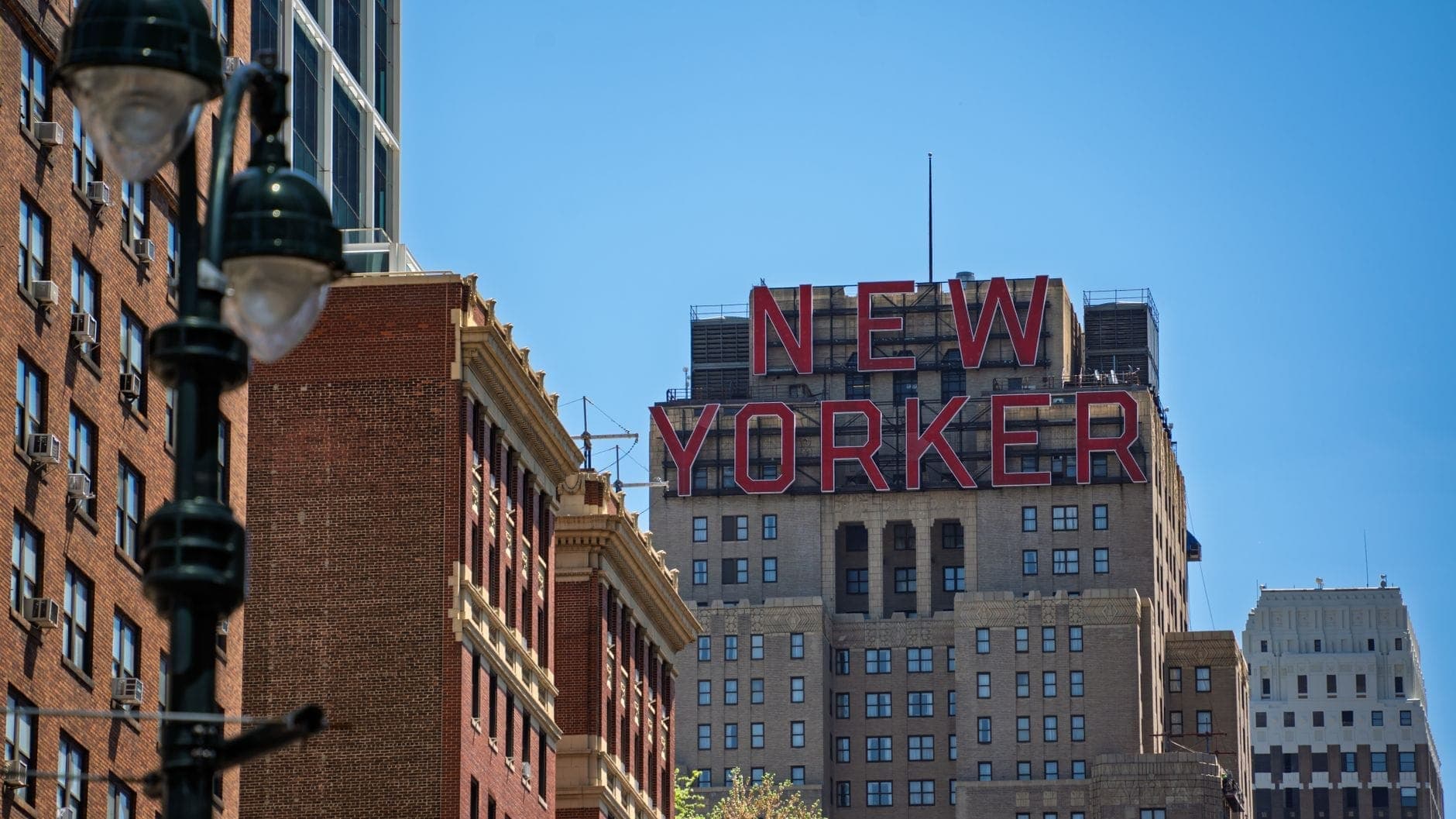 อาคารโรงแรม New Yorker อันโดดเด่นพร้อมป้ายสีแดงขนาดใหญ่ ล้อมรอบด้วยสถาปัตยกรรมของ Midtown Manhattan ในวันที่อากาศแจ่มใส