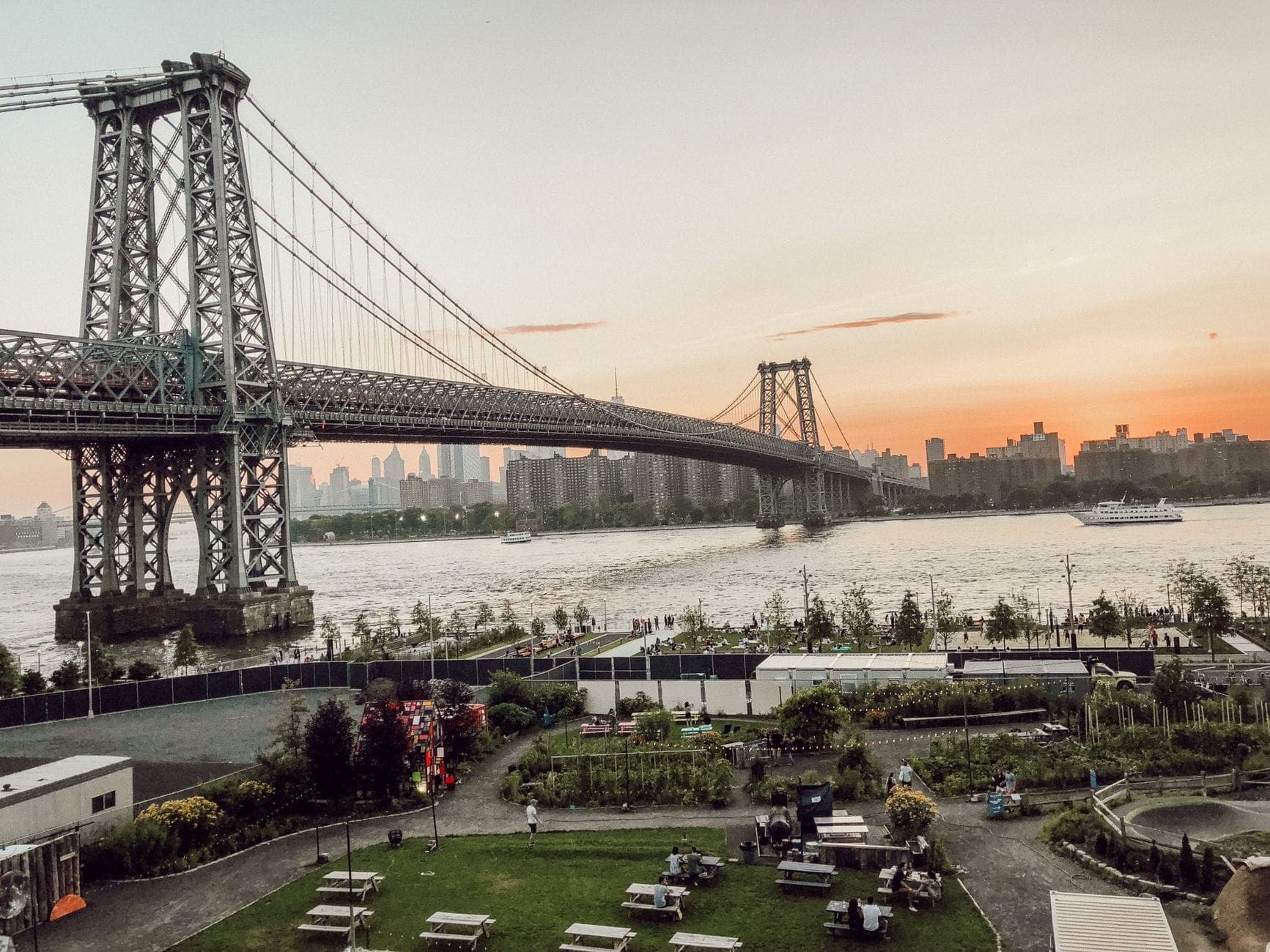 วิวของ Williamsburg Bridge ที่ทอดข้าม East River โดยมีเส้นขอบฟ้าเป็นฉากหลัง และผู้คนกำลังพักผ่อนในสวนสาธารณะริมน้ำสีเขียวใน Brooklyn