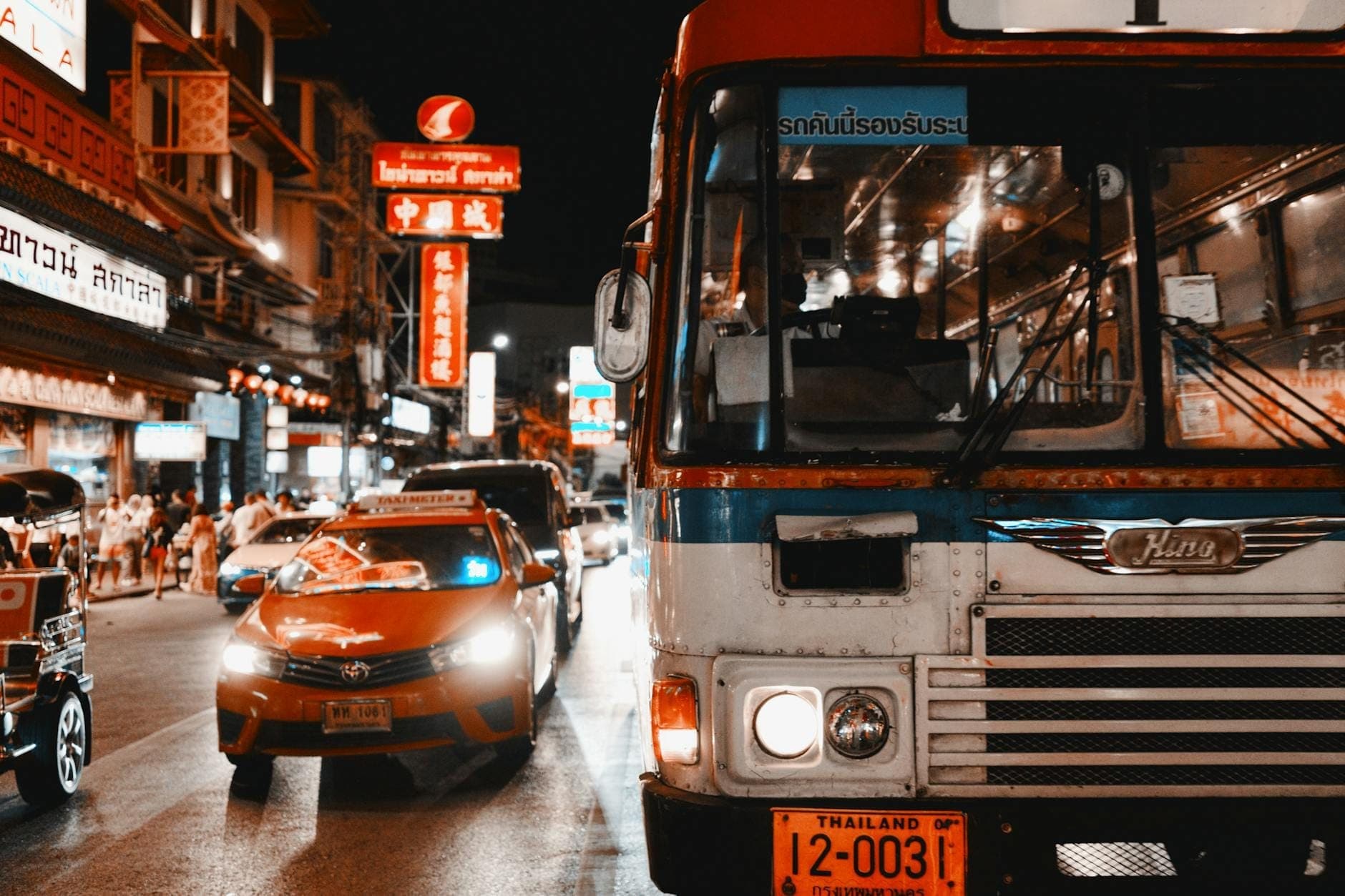 Eksena sa kalye ng gabi sa Thailand na may taksi, tuk tuk, at bus sa gitna ng mga neon na karatula, na kumukuha ng tipikal na lokal na transportasyon.