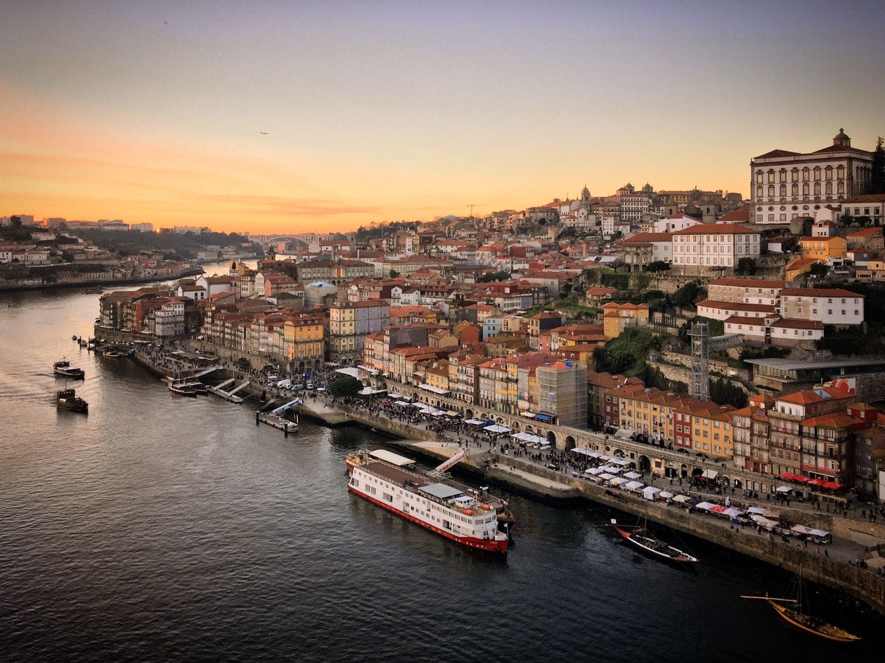มุมมองทางอากาศของย่าน Ribeira ในเมือง Porto พร้อมอาคารสีสันสดใสริมแม่น้ำ ทางเดินเลียบแม่น้ำ Douro เรือ และท้องฟ้ายามอาทิตย์อัสดงเหนือเมือง