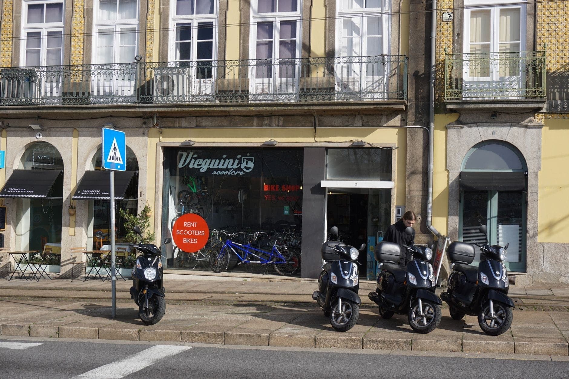 แถวของสกู๊ตเตอร์ที่จอดอยู่หน้าร้านค้าท้องถิ่นและคาเฟ่ในเมือง Porto พร้อมสถาปัตยกรรมโปรตุเกสคลาสสิกสีเหลืองและระเบียงด้านบน