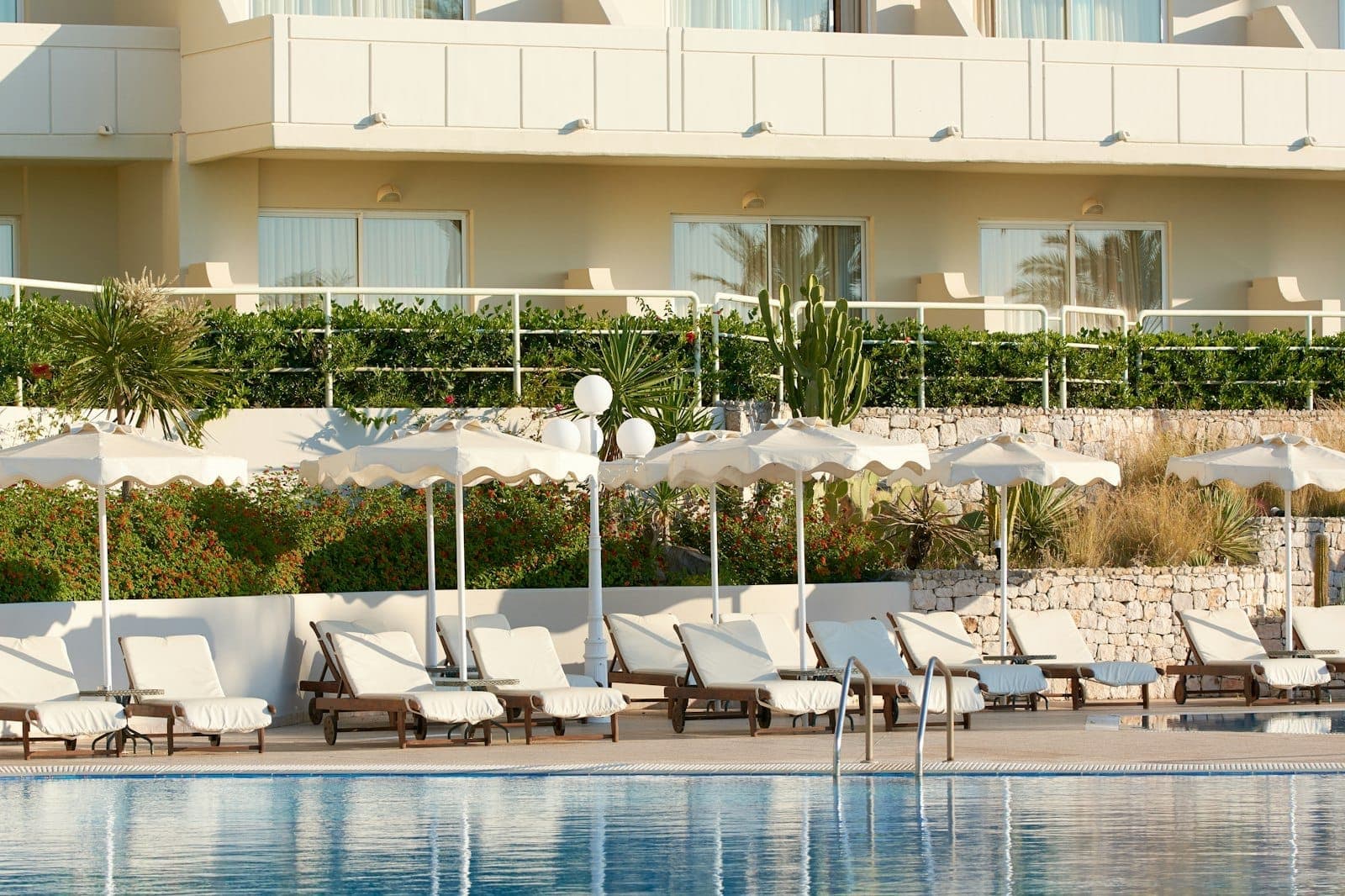 Hôtel moderne à Rhodes avec balcons donnant sur une piscine aménagée, transats, parasols blancs et plantes méditerranéennes par une journée ensoleillée.