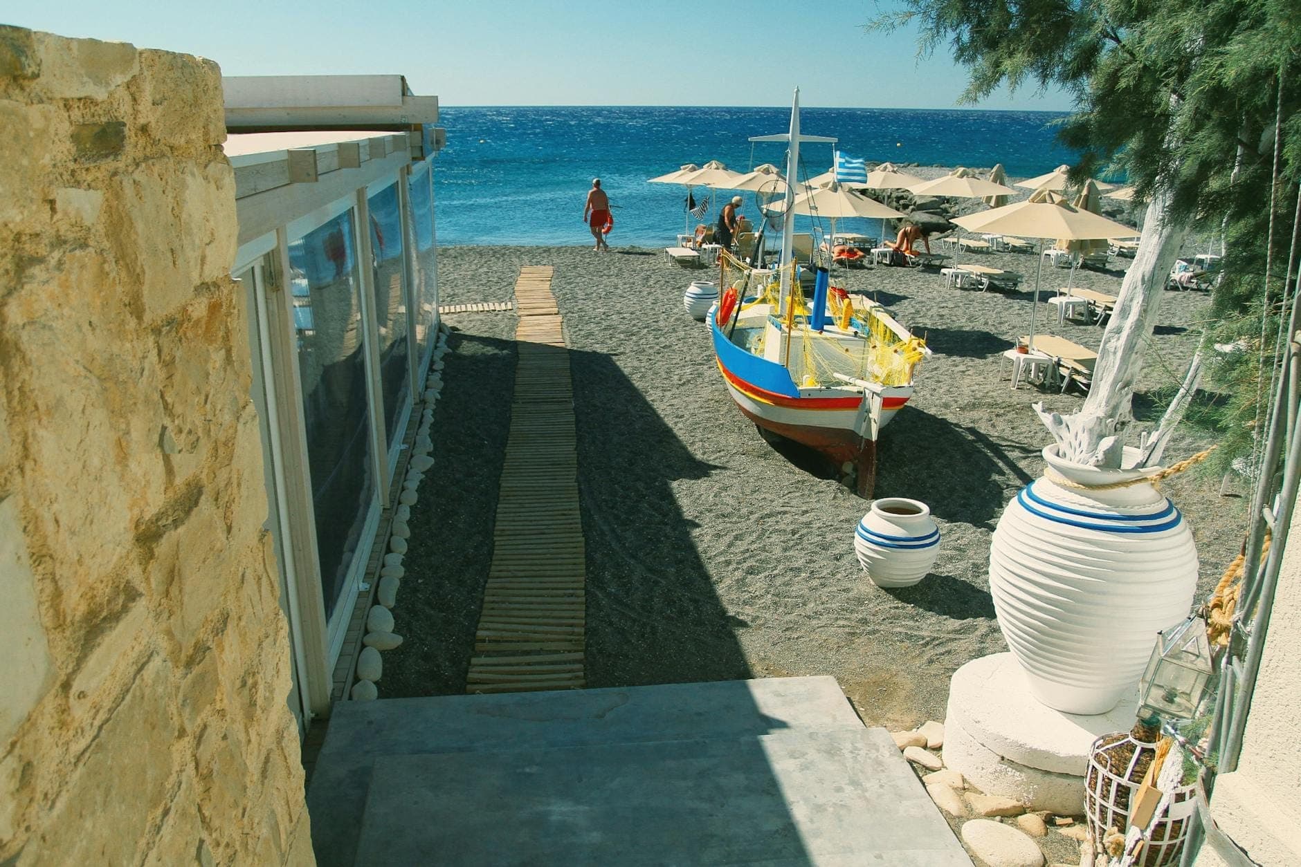 Sandy beach scene with a boardwalk, colorful fishing boat, sun umbrellas, lounge chairs, and blue sea in a Santorini beach town.