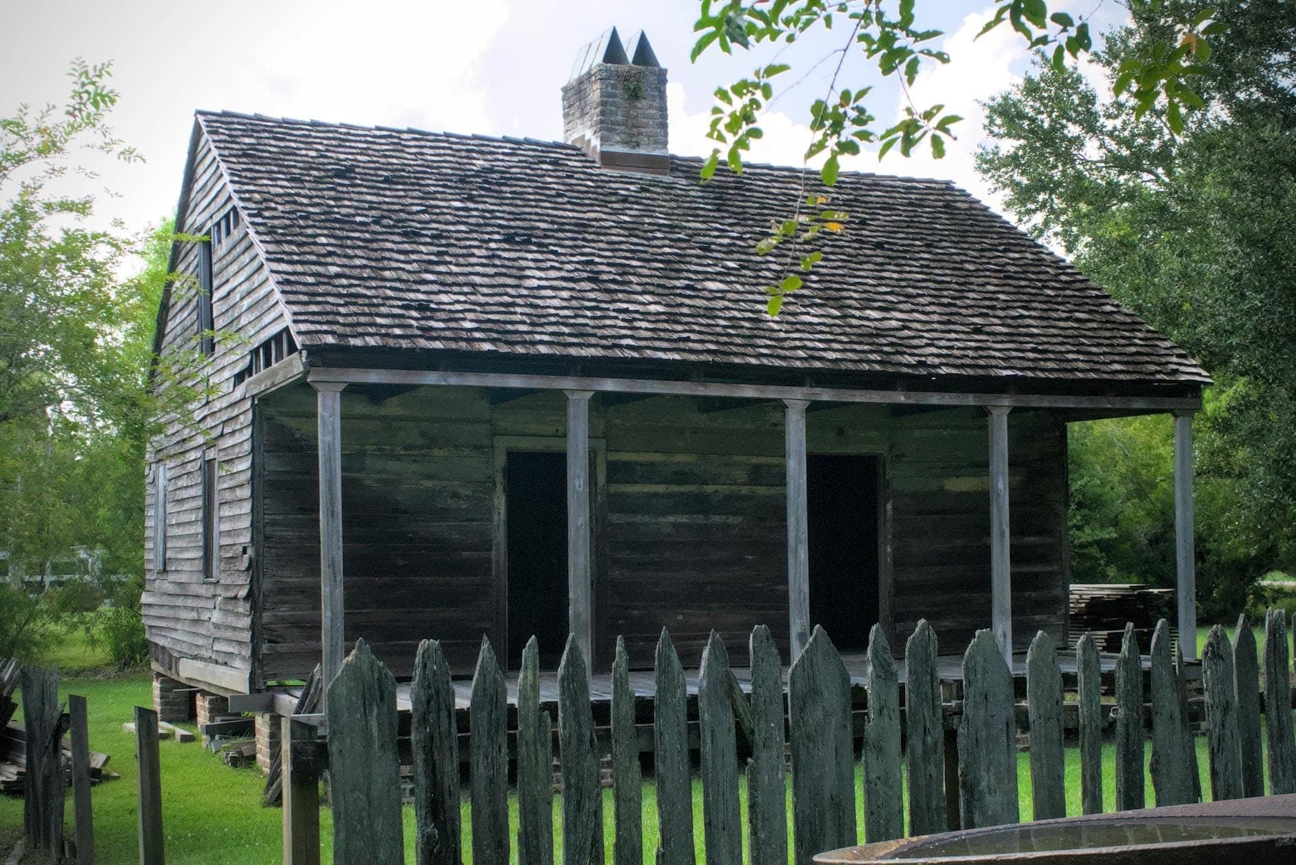 A weathered wooden slave cabin behind a picket fence, surrounded by green grass and trees under a partly cloudy sky.