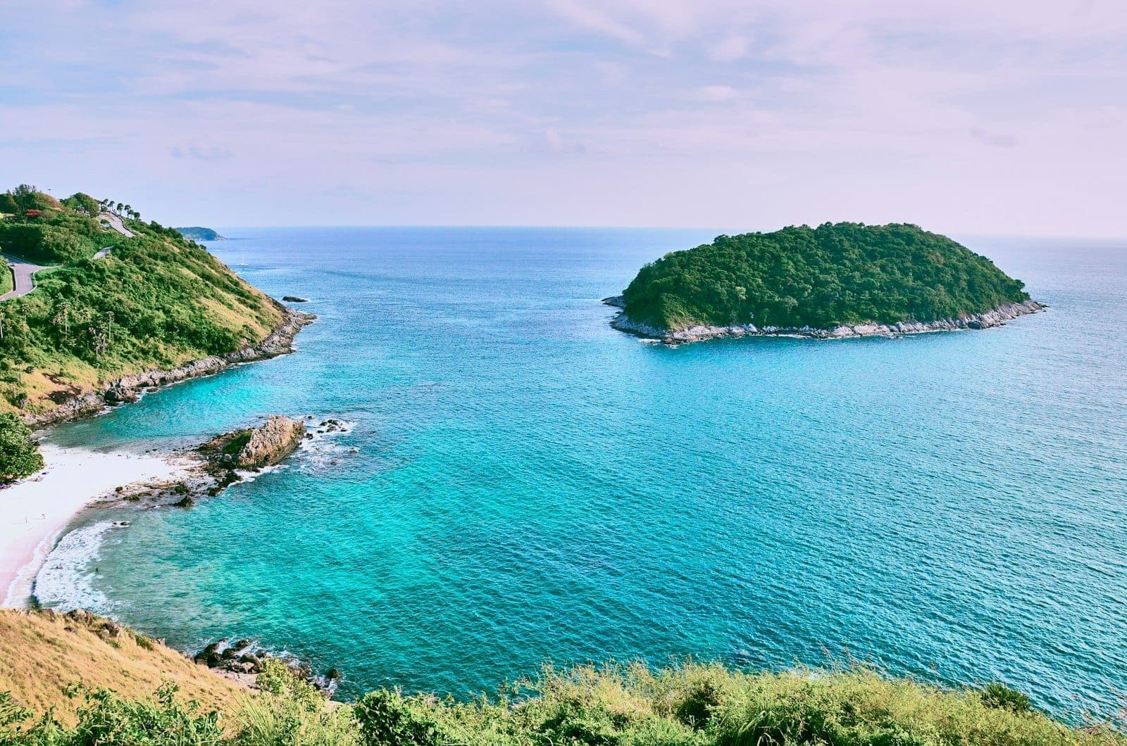 Expansive daytime view from Windmill Viewpoint Phuket overlooking turquoise water, lush hills, white sand beach, and a small green island in the Andaman Sea.