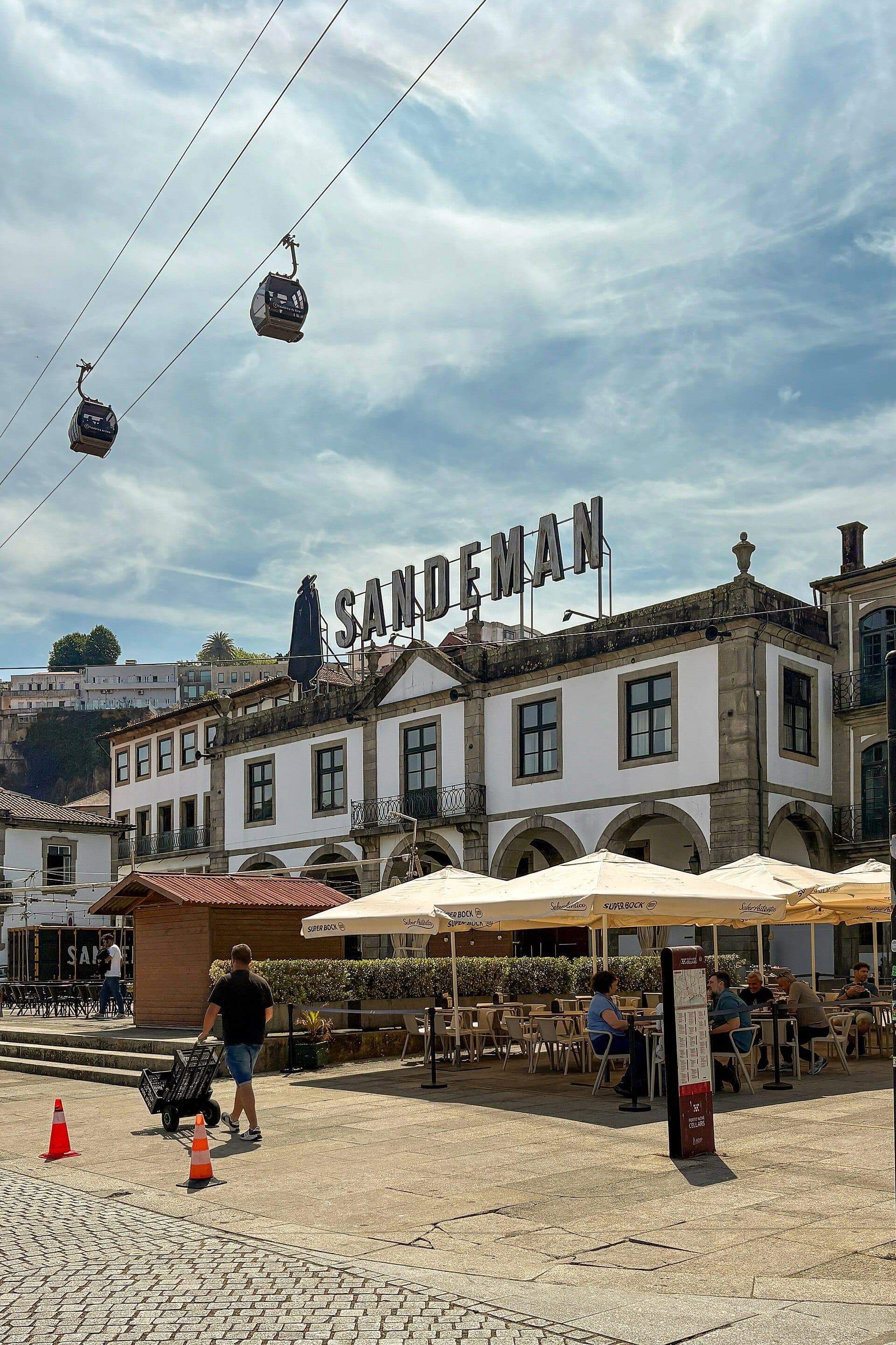 Area pranzo all'aperto con commensali sotto ombrelloni bianchi davanti all'edificio Sandeman al WOW Porto, con funivie che passano in alto.