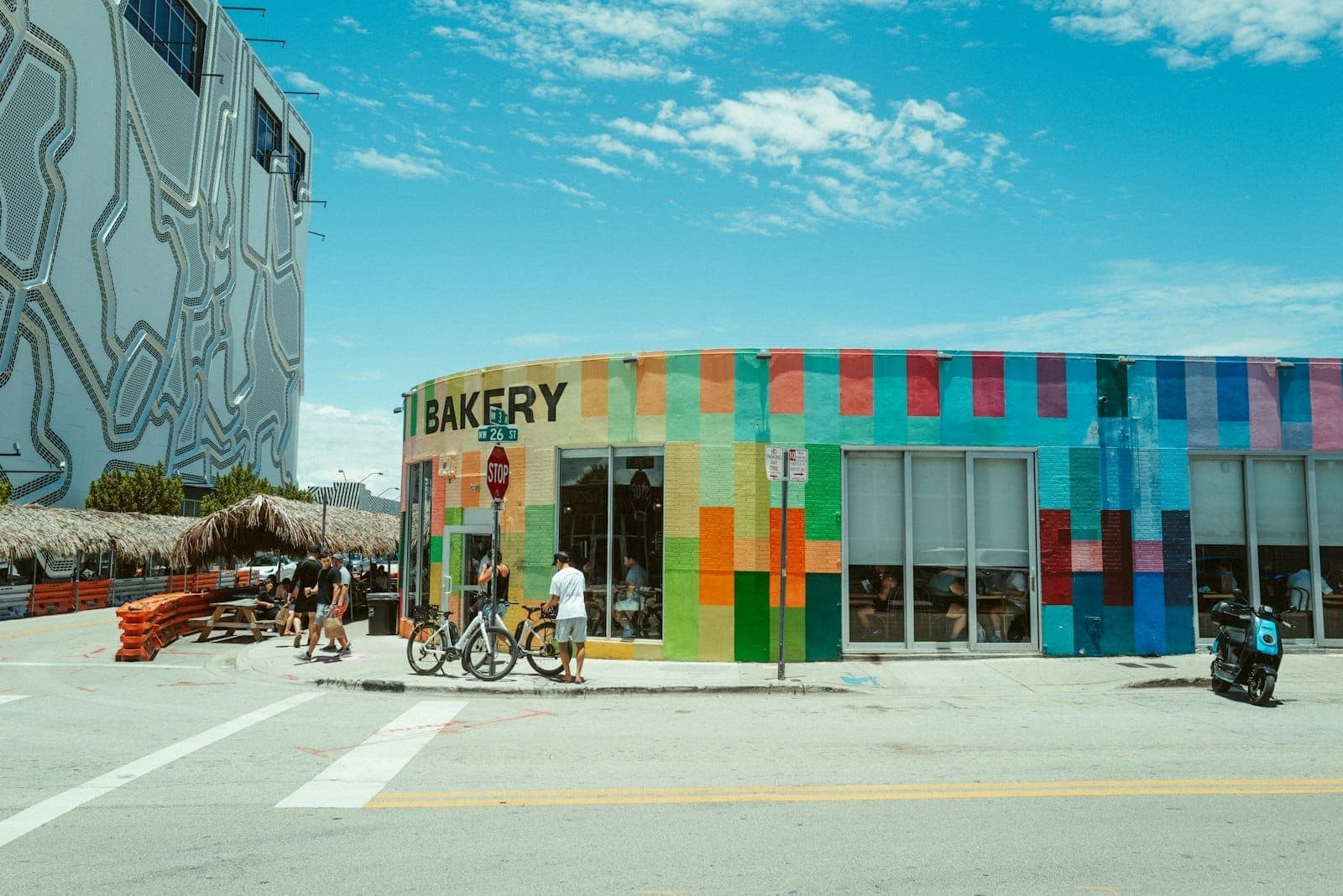 A colorful, modern bakery building with vibrant street art, people walking and biking, under a bright blue sky in Wynwood, Miami.